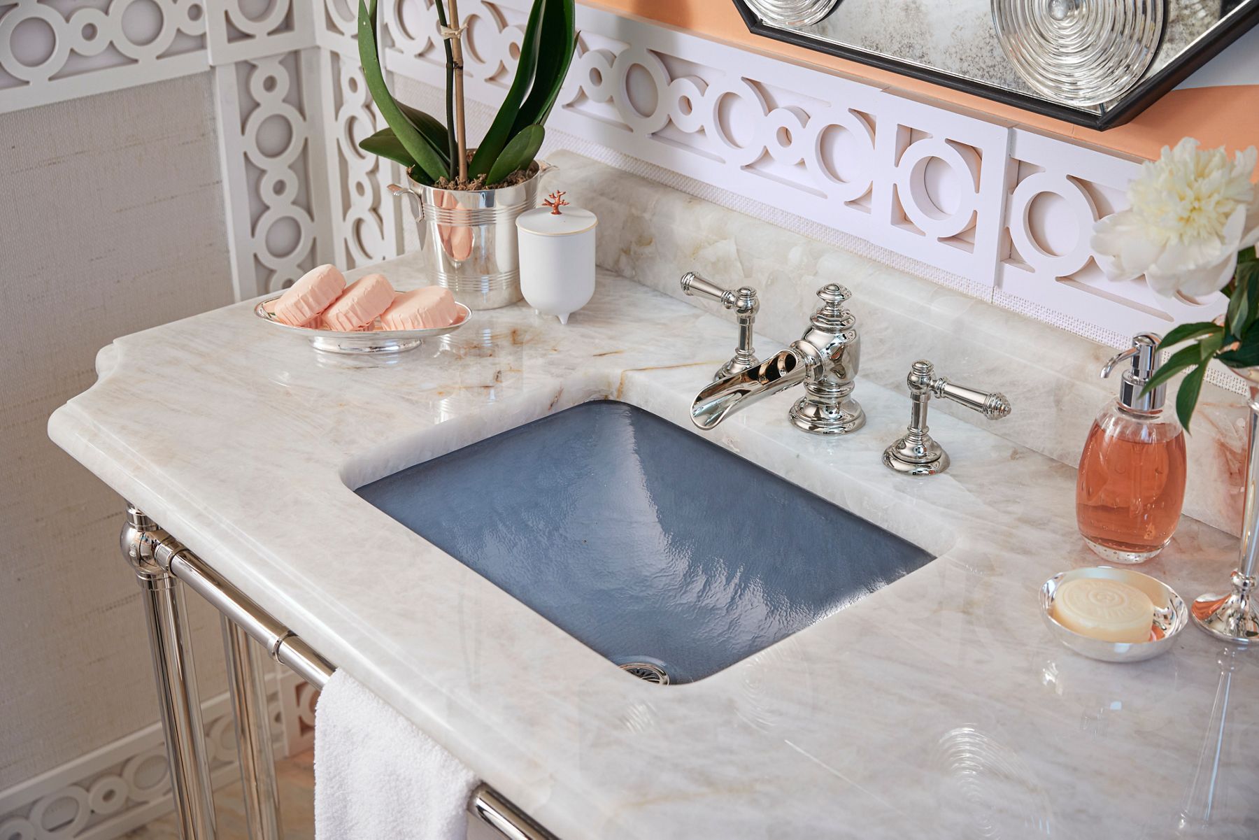 Marble bathroom vanity with a soft blue sink basin, polished nickel Kohler faucet with cross handles, decorative geometric wall detailing, and styled accessories including soap dispensers, candles, and flowers.