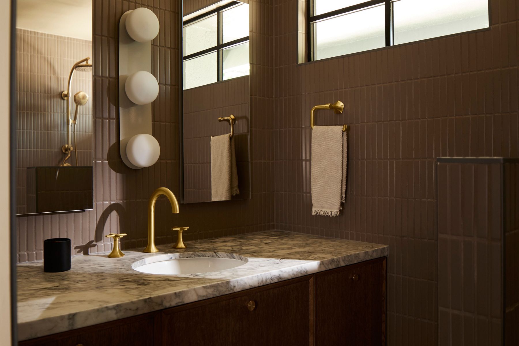 Contemporary bathroom vanity with marble countertop, brass faucet, dark wood cabinetry, and brown tiled walls illuminated by globe sconces.