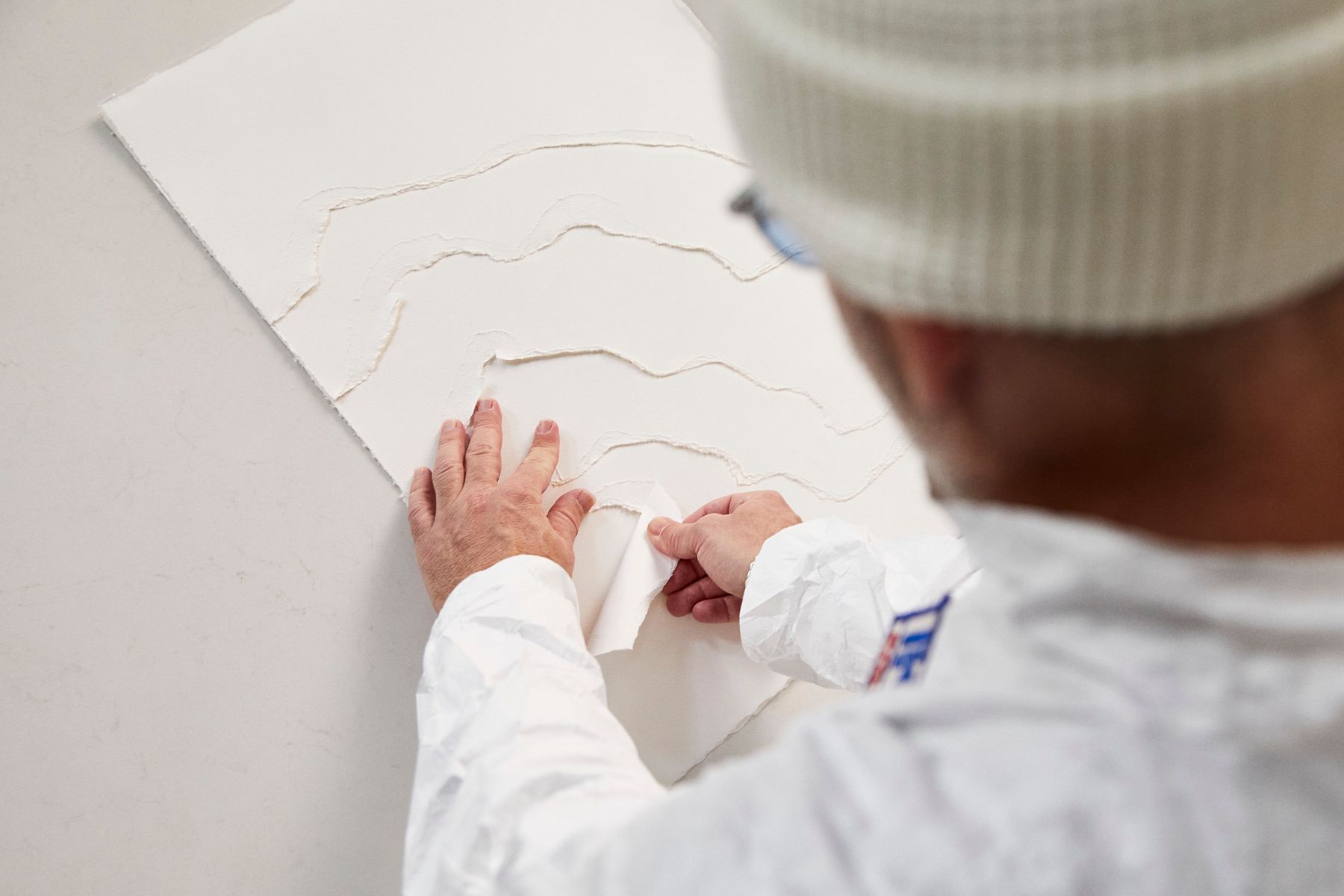 A person wearing a white jacket and a white beanie is carefully peeling away layers from a textured, white paper-like material on a flat surface, revealing underlying shapes and patterns.