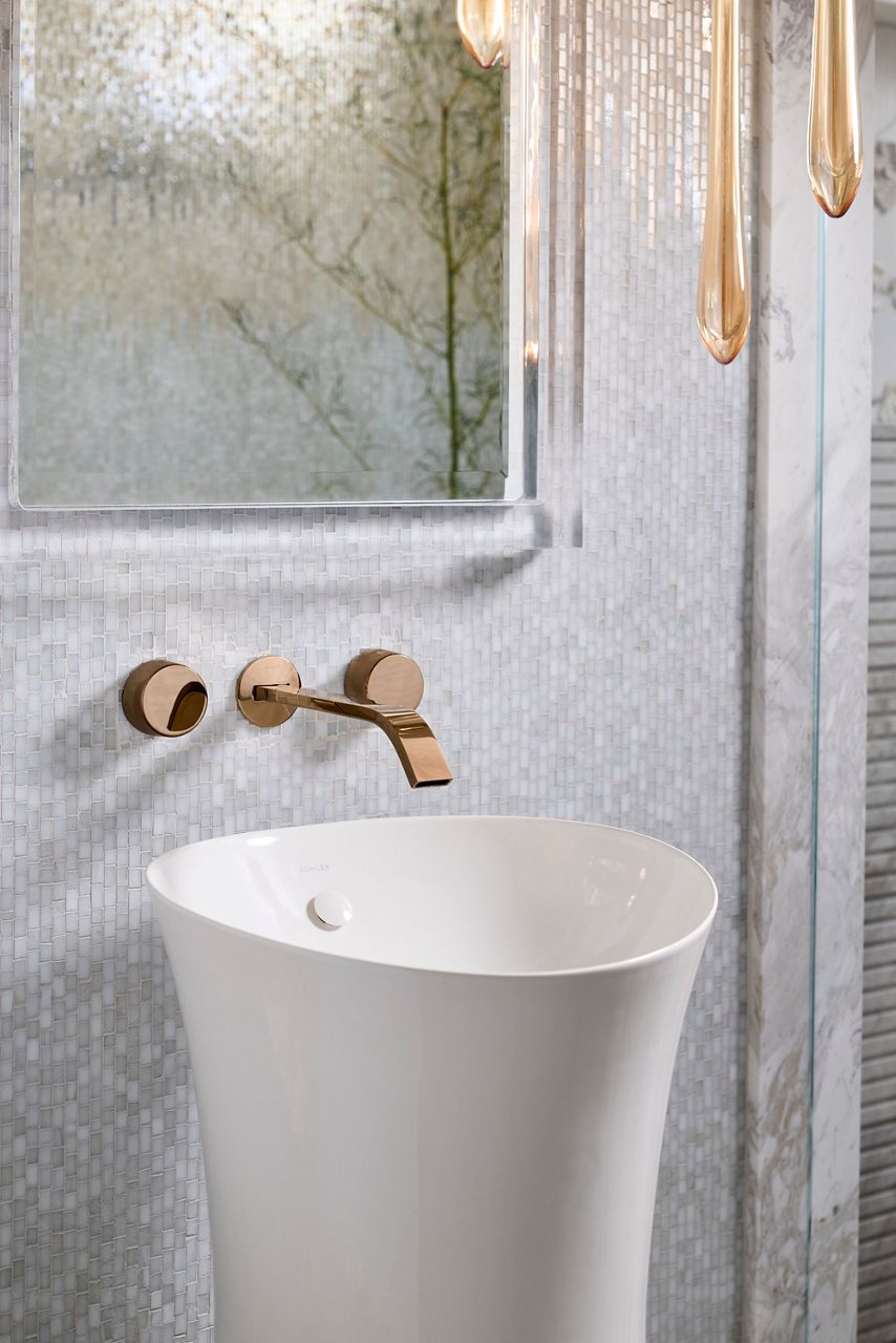 White pedestal sink with wall-mounted brass faucet and textured glass wall backdrop in a modern bathroom setting.