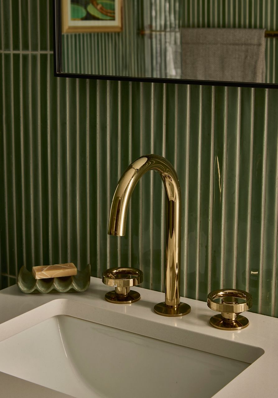 Close-up of a polished brass widespread bathroom faucet mounted on a white sink against dark green vertically striped tile walls.