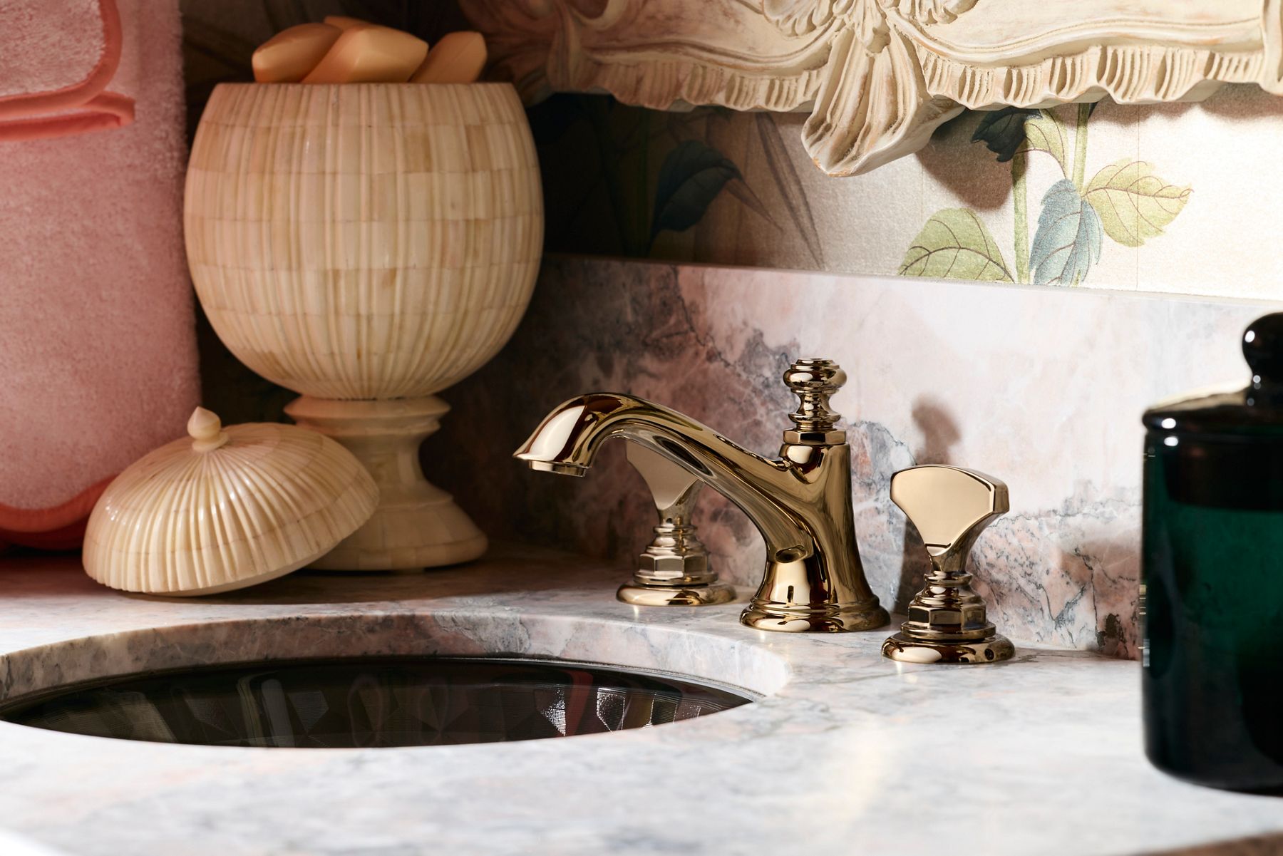 Traditional brass bathroom faucet beside a dark vessel sink with decorative accessories and floral wallpaper accents.