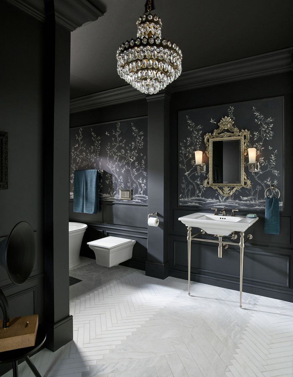 A Memoirs console table sink, toilet, and freestanding bath in a bathroom with dark gray walls with ornate white plant life drawings and an ornate gold-framed mirror.