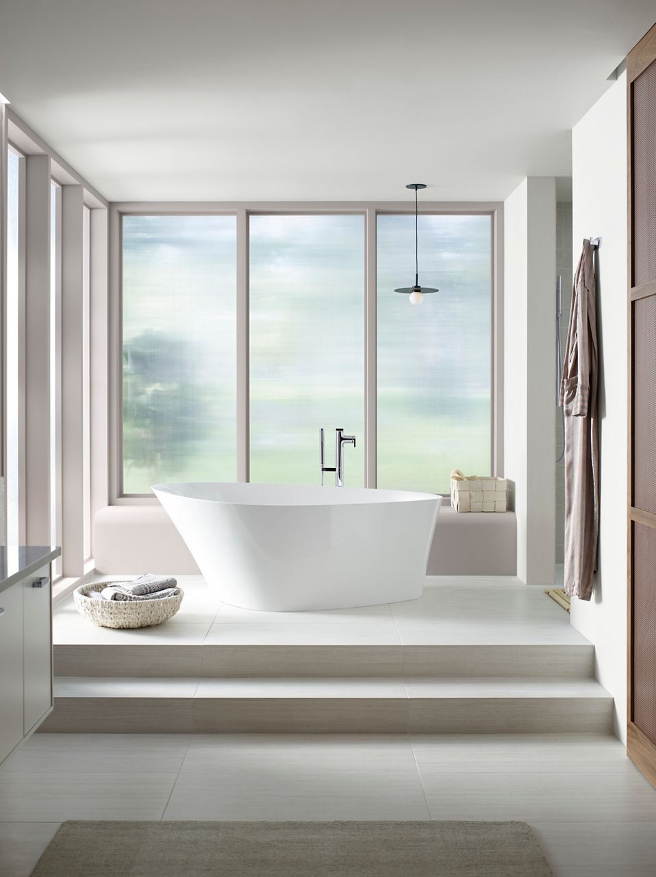 A Veil freestanding bath set in a large room on a raised floor in a room with a neutral color scheme with a large semi-opaque window looking out over a blurred nature scene.