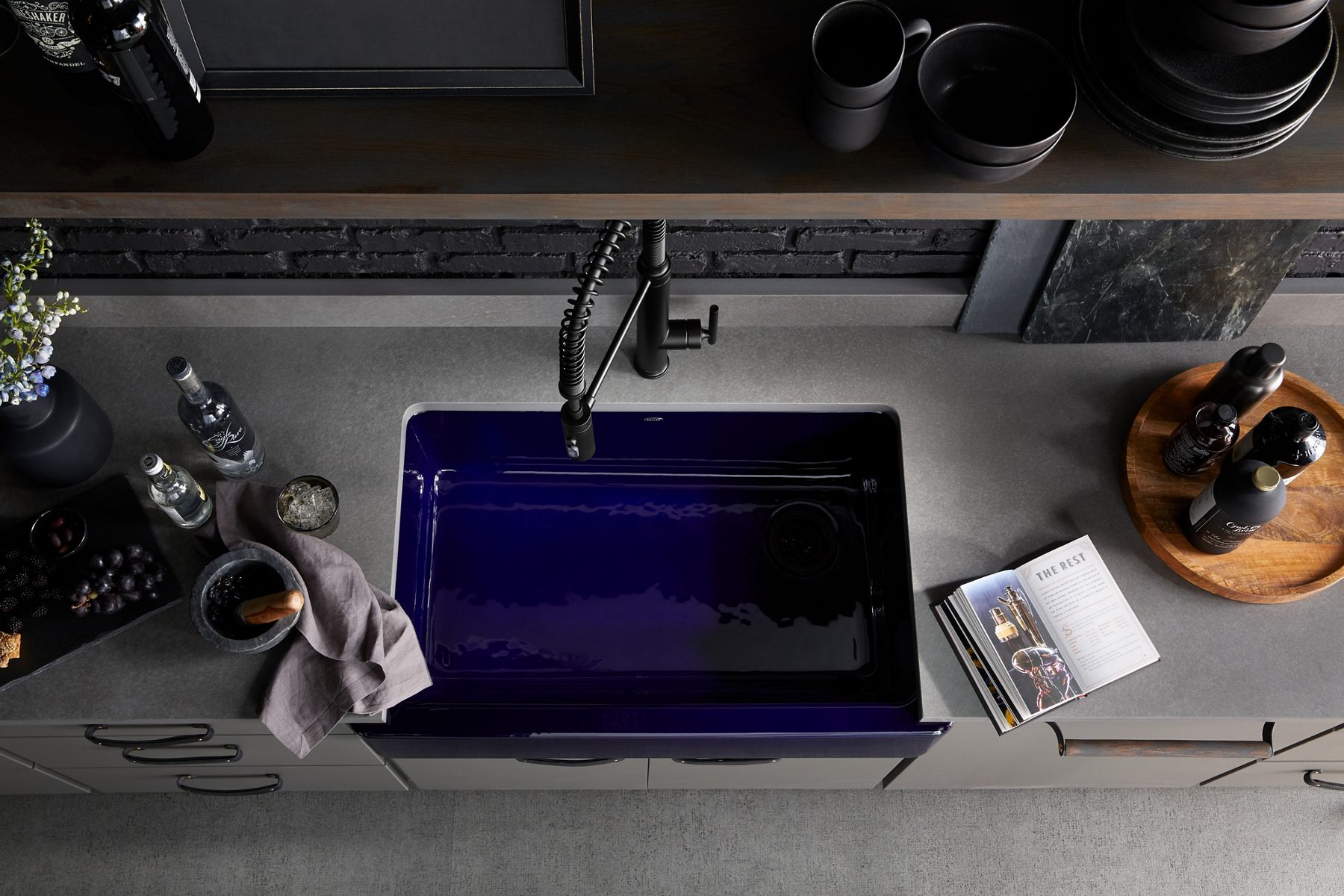 Aerial view of Whitehaven self-trimming, under-mount kitchen sink with tall apron in Indigo Blue paired with a semi-professional kitchen faucet finished in Matte Black installed in a grey-toned, modern kitchen with open shelving. 