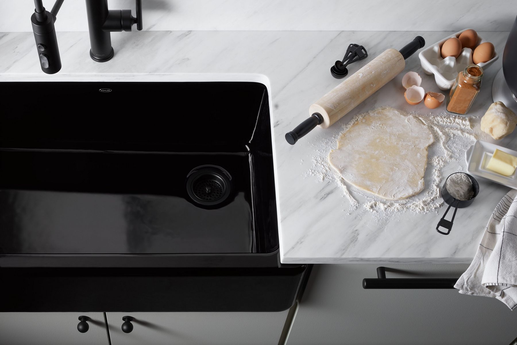 Overhead view of a black enameled cast iron apron front sink set in a stone counter top with a rolling pin, dough, butter, egg shells and other baking items arranged on the side.