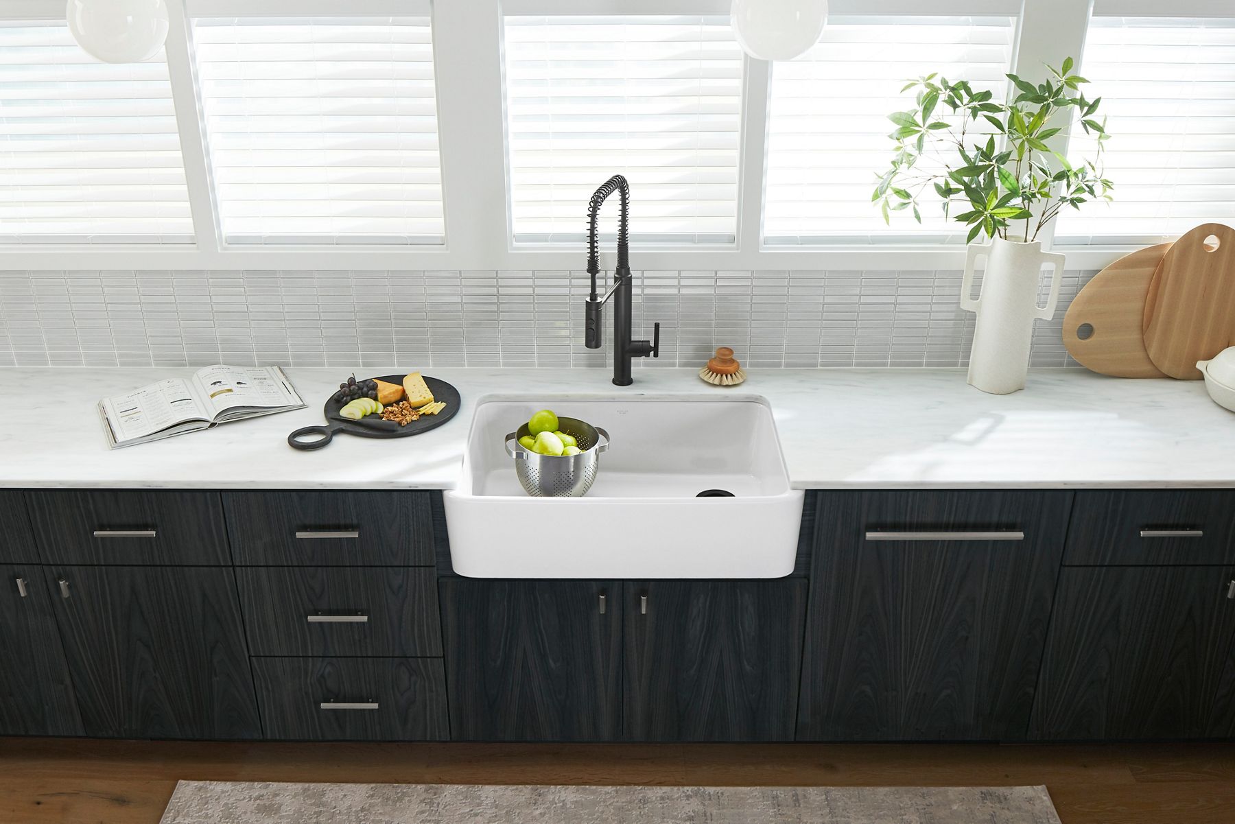 White countertop with deep farmhouse sink, black industrial-style faucet, and colander of green apples.
