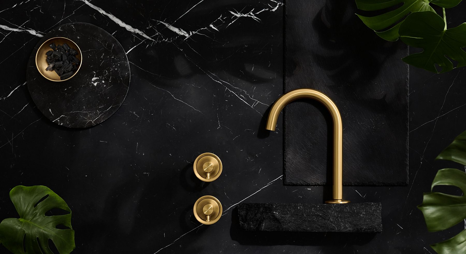 A vibrant brushed moderne brass faucet and handles laying down on a dark gray stone surface with a brass bowl nearby.