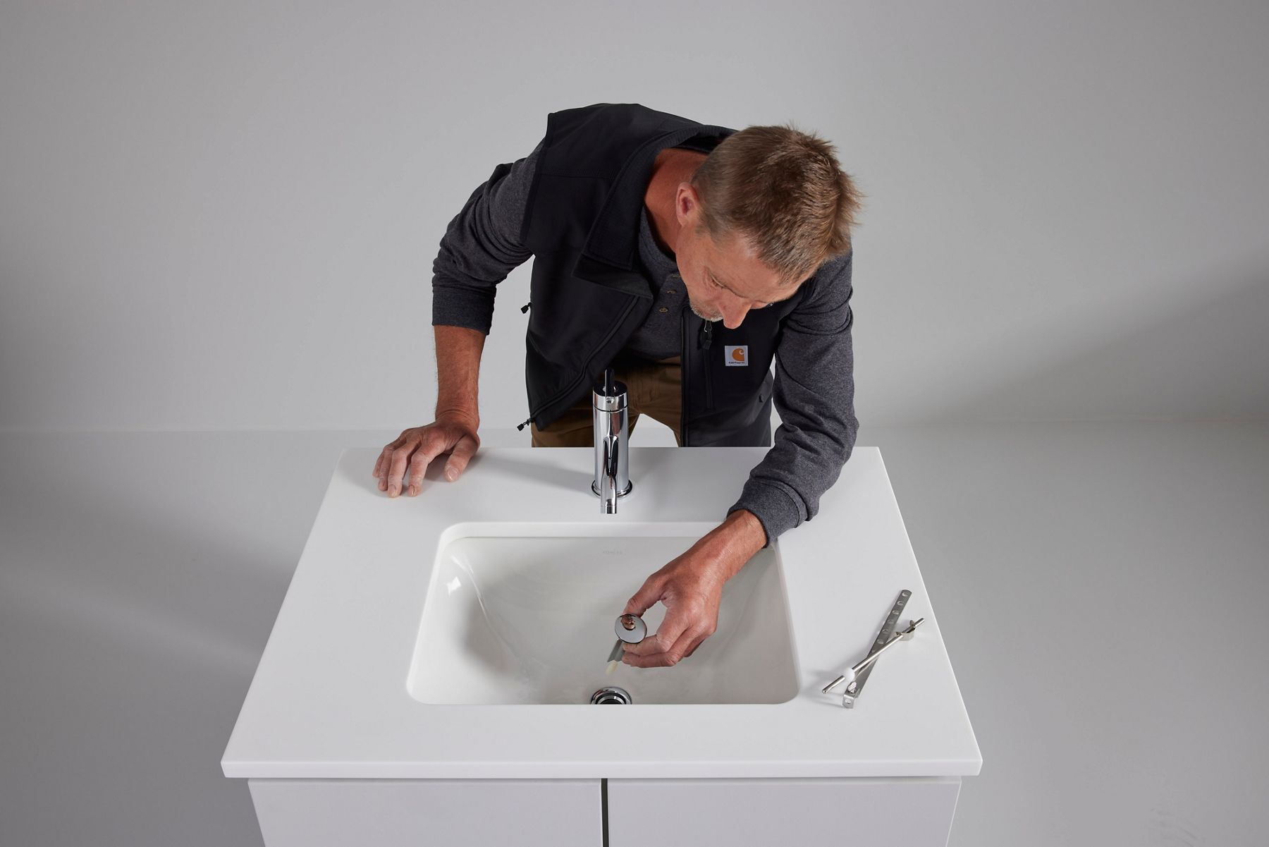 Person installing or adjusting a bathroom sink drain assembly in a white vanity sink, viewed from above, with installation tools placed beside the basin.