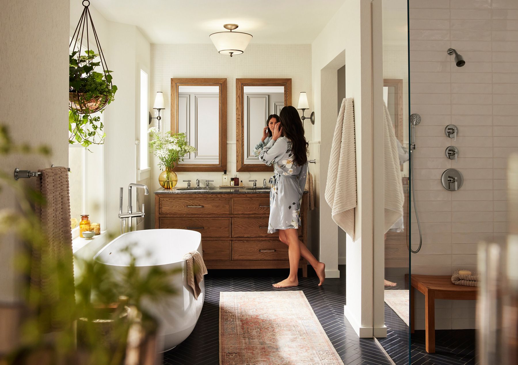 Elegant bathroom interior featuring a freestanding white bathtub with a towel draped over the edge, positioned next to a wooden double vanity with two large framed mirrors above it. The room is softly lit by wall sconces on either side of the mirrors and a central ceiling light. A potted plant in a golden vase and fresh flowers add a touch of greenery, while a neutral-toned rug lies on the dark herringbone-patterned floor. Light, cream-colored walls and ample natural light create a serene and cozy atmosphere.