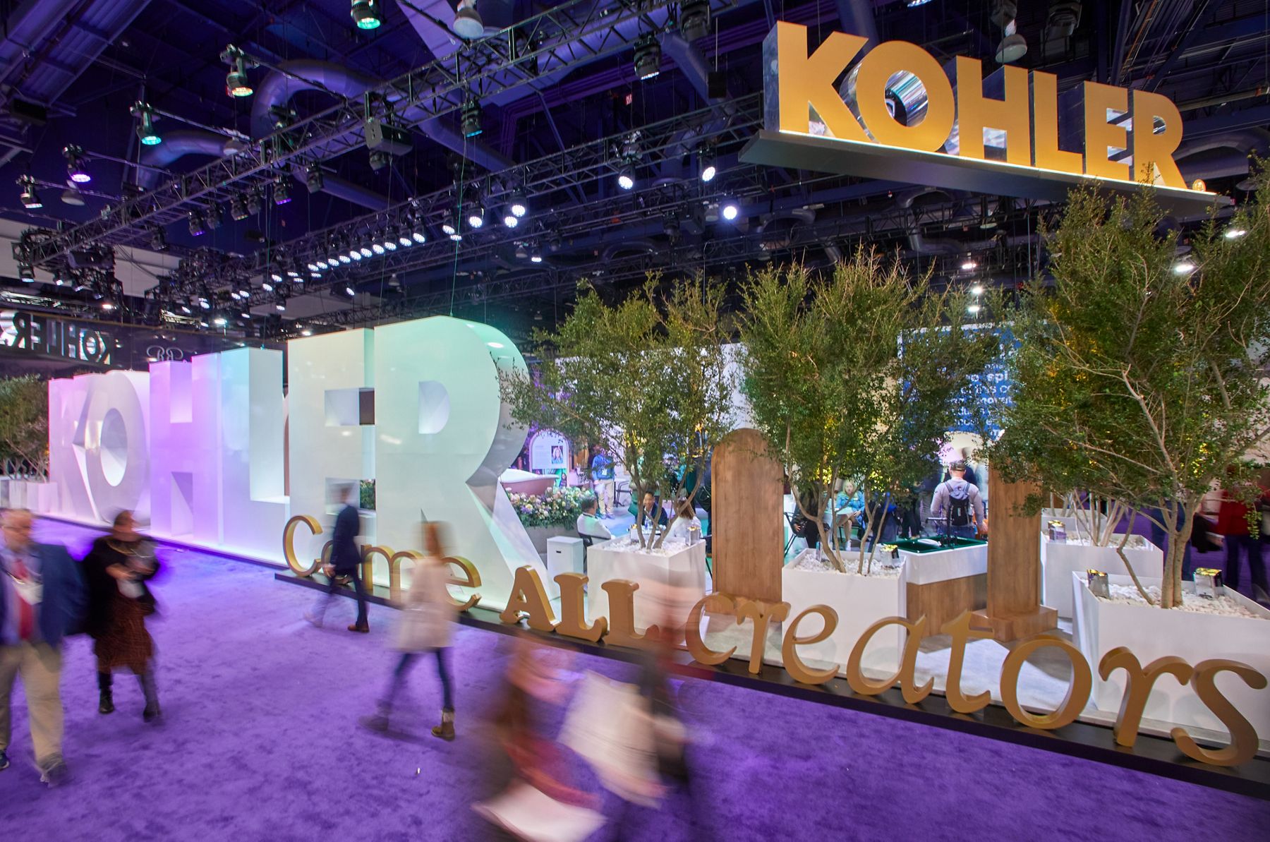Un kiosque de salon commercial Kohler avec le mot KOHLER en gros lettrage tridimensionnel blanc, environ dix pieds de haut, et les mots Come All Creators en plus petits lettrage tridimensionnel en trois faces différentes, environ trois pieds de haut, avec plusieurs arbres et des affichages derrière eux alors que les gens passent dans un flou à vitesse d’obturation lente.