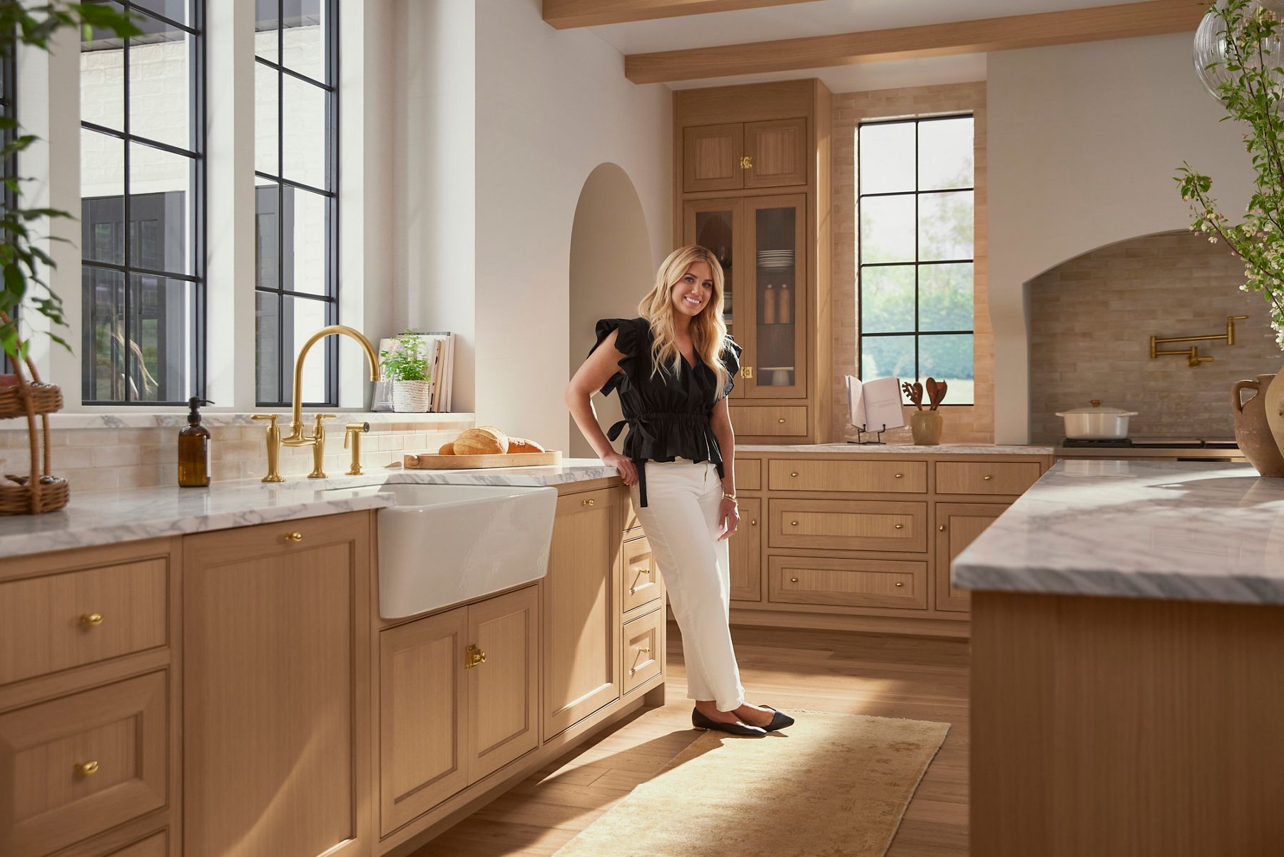 Designer Shea McGee standing in a neutral tone kitchen next to a brushed brass faucet she designed.