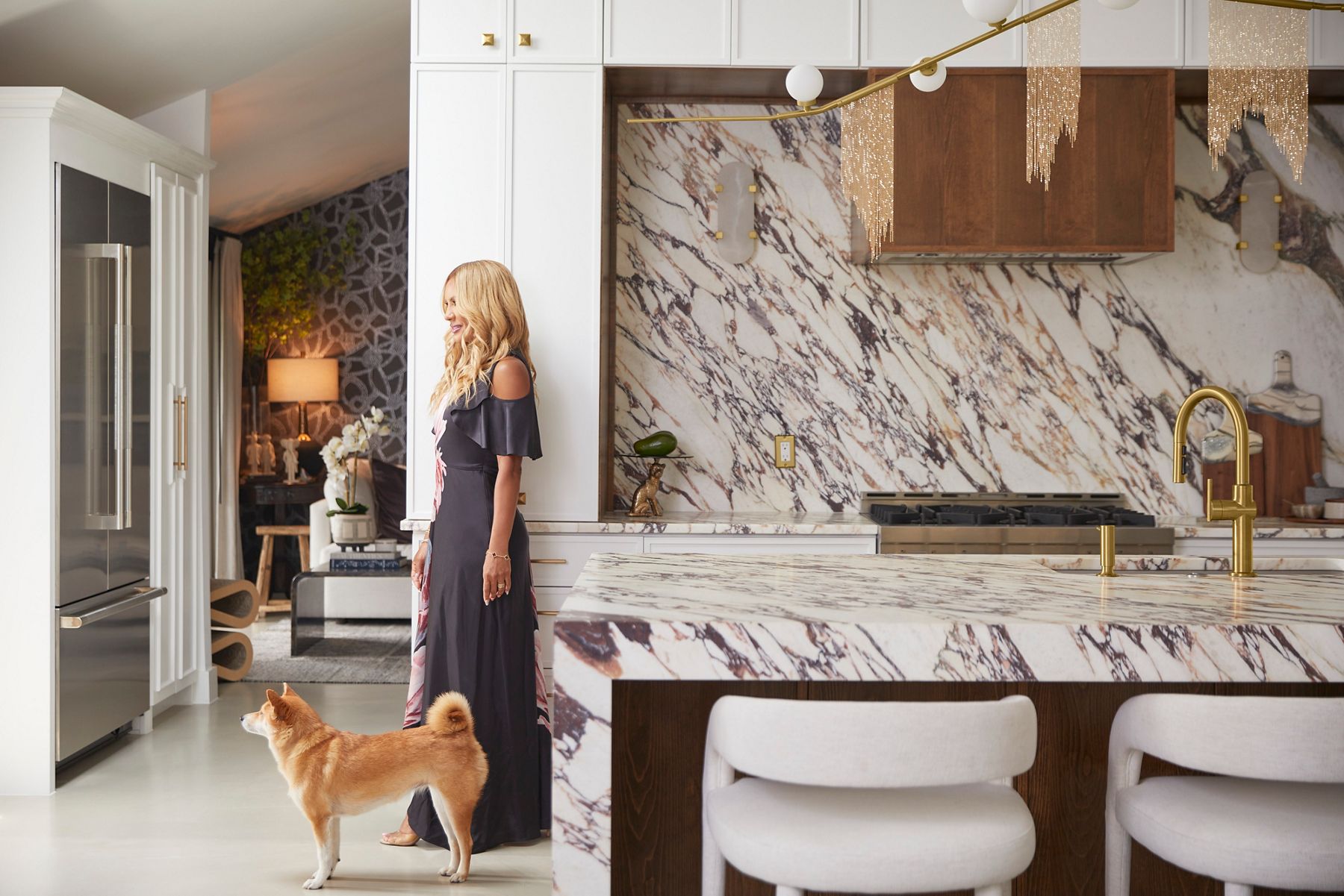 Wide image of Nikki Chu standing in her kitchen with her dog at her feet. They are both looking off to the left and leaning against the white and purple-veined marble counter top, which matches the large kitchen island counter top. In the distance you can see Nikki's living room and in the foreground, you can see her kitchen faucet, a Kohler Crue faucet in brass.