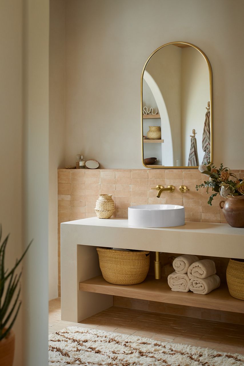 A bathroom vanity area in warm neutral tones with a wall-mount brushed brass faucet over a round white vessel sink.