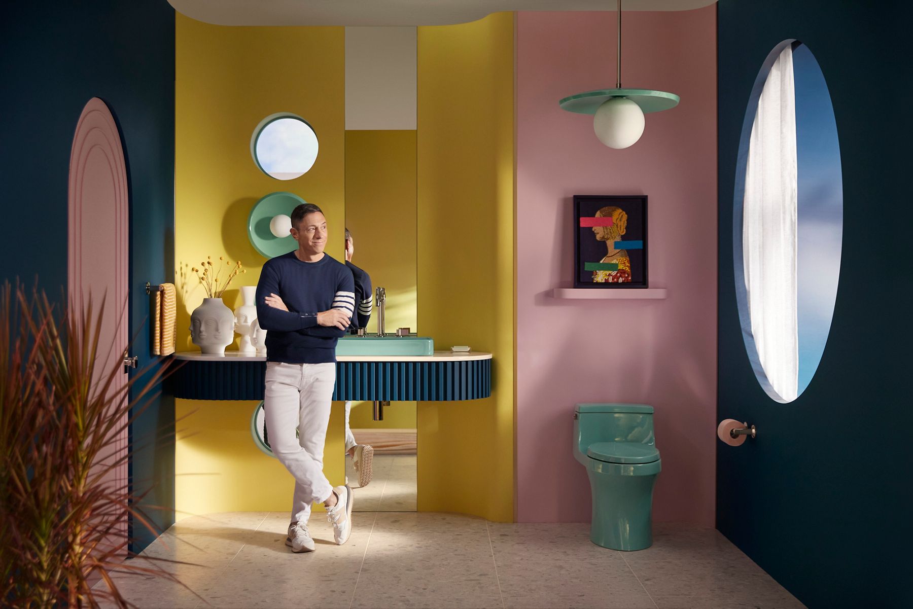  Image of acclaimed designer and author Jonathan Adler standing in the bathroom he designed with Kohler's new Heritage Color products in the vintage color called Spring Green. He's leaning against the vanity featuring a colorful vessel sink and looking out the sunny window. 