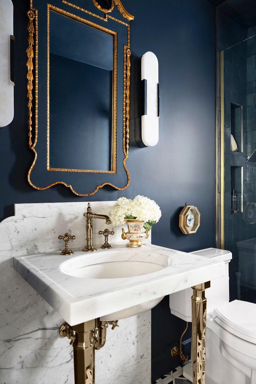 A landscape image of an elegant guest bathroom featuring deep blue walls, a white marble countertop with polished metal fixtures, a decorative vase with white hydrangeas, and a gold-framed octagonal mirror.