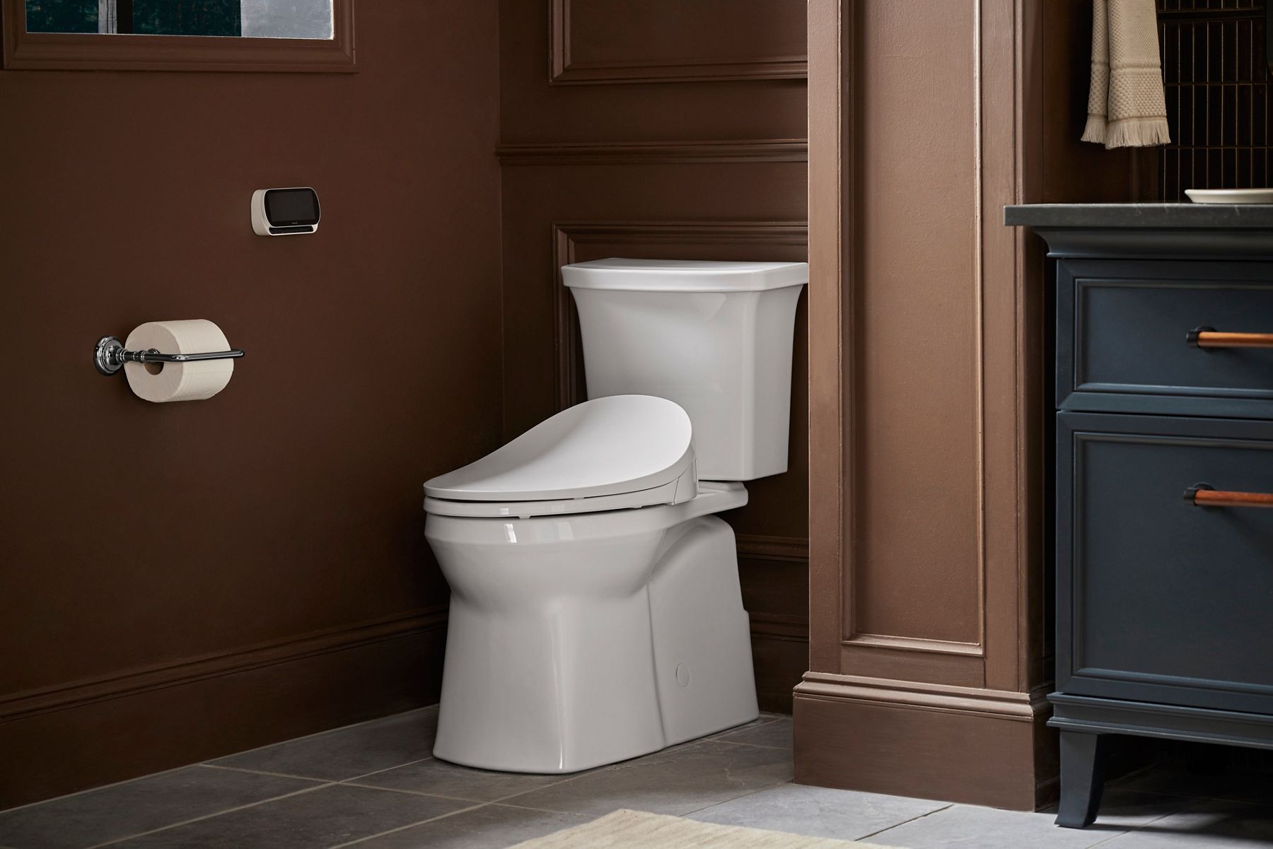 White modern toilet with closed lid in a bathroom with brown wall and toilet paper holder mounted on wall.