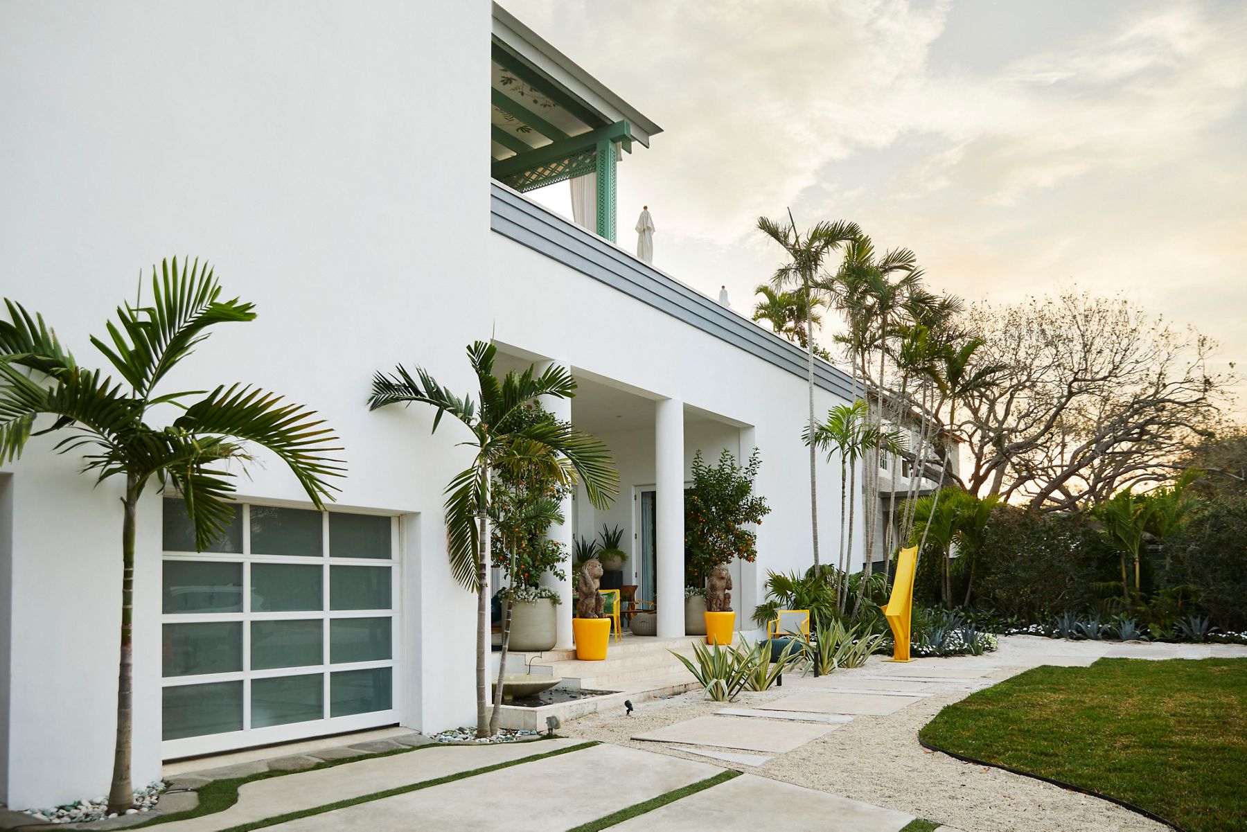 Exterior of the Kips Bay Palm Beach show house with a white building, bright yellow sculptures and planters, and abundant tropical plants.