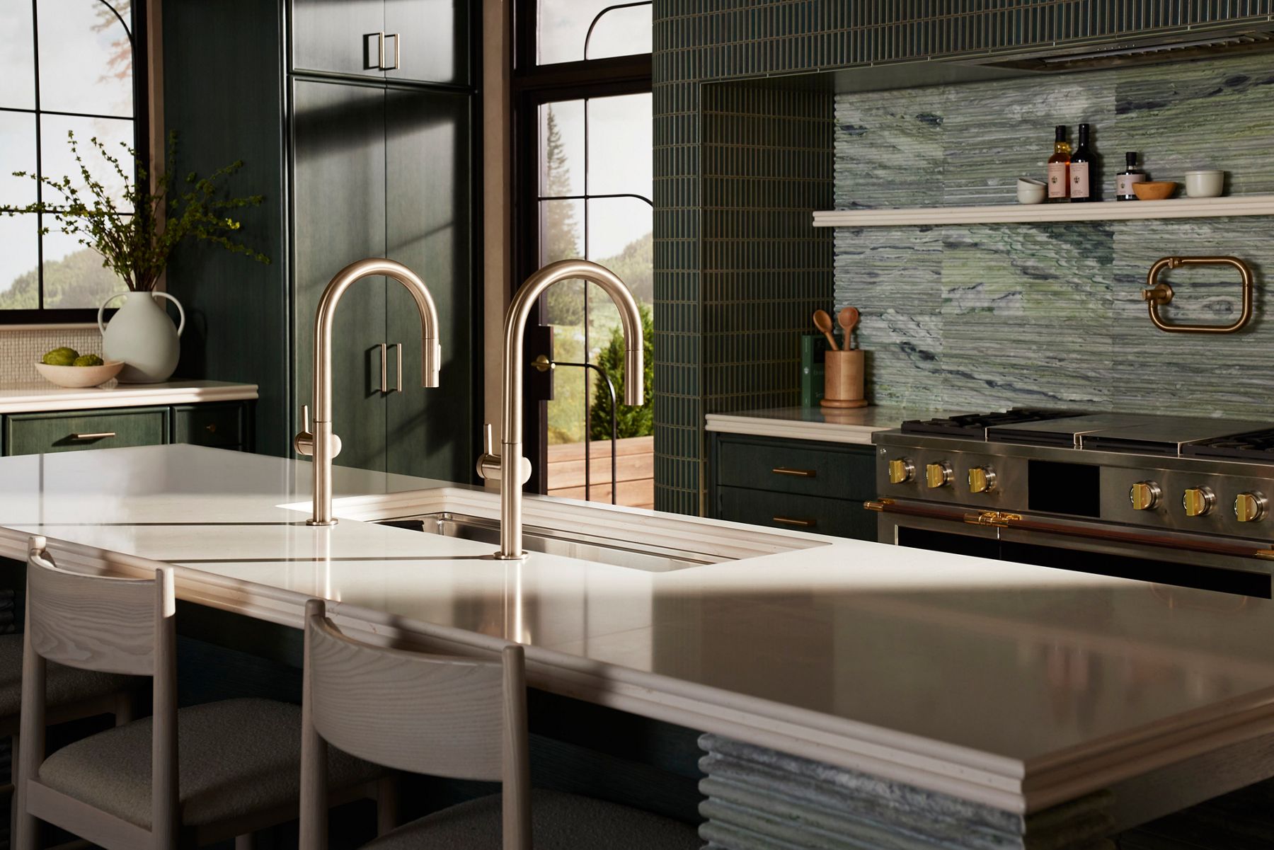 Contemporary kitchen with warm metallic bridge faucets, white island countertop, dark cabinetry, and a textured stone backsplash under natural light.