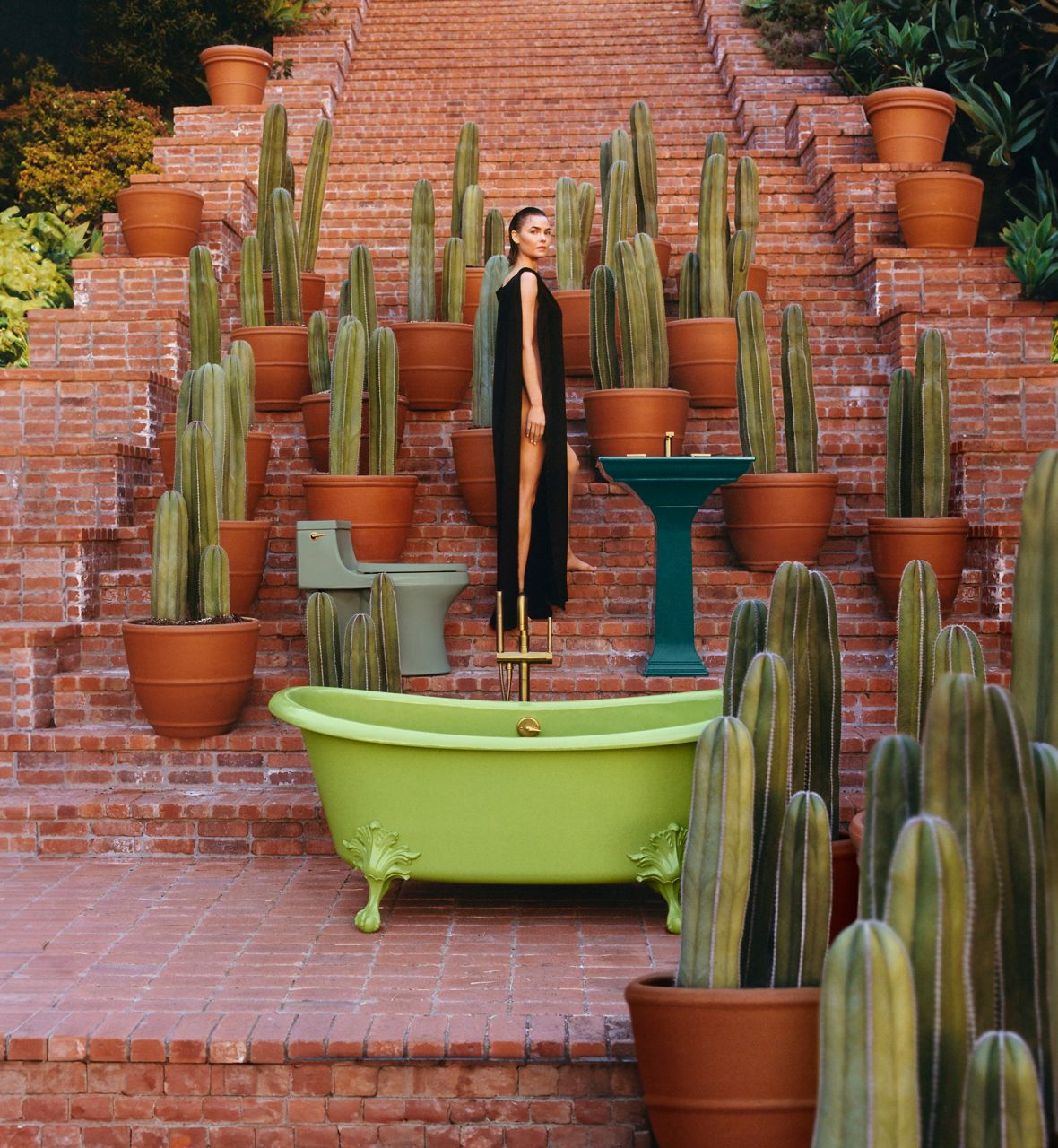A wide shot lifestyle image taken on a long brick stairwell. A woman is standing at the center in a black dress and positioned at her side. There are potted cacti around her and a bright green claw tub at the front of the image. There's a teal kitchen sink on her right side and a pale green toilet behind her.