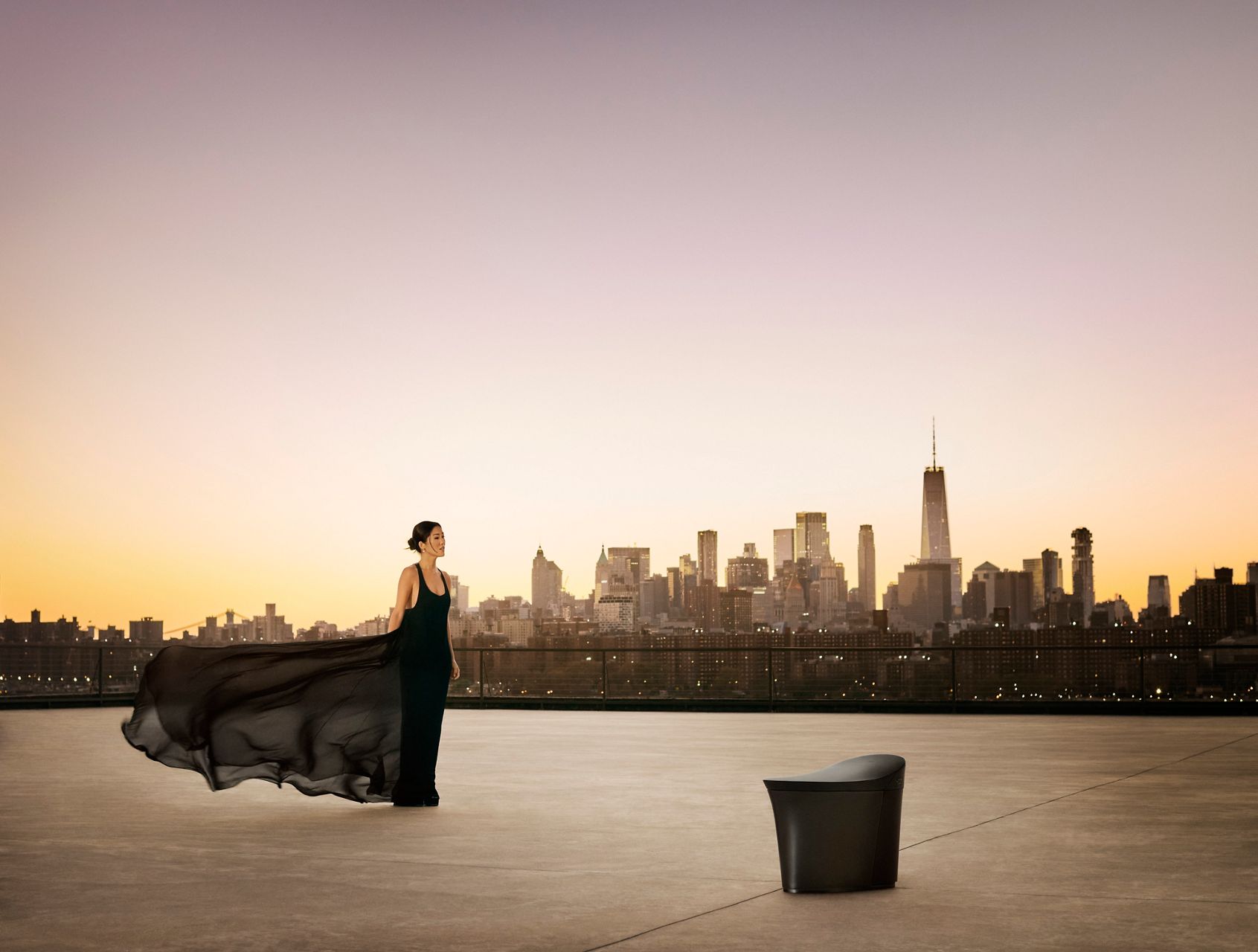 Laura Kim stands in a flowing black chiffon dress, accentuating the curved design of the Veil smart toilet against the New York City skyline.