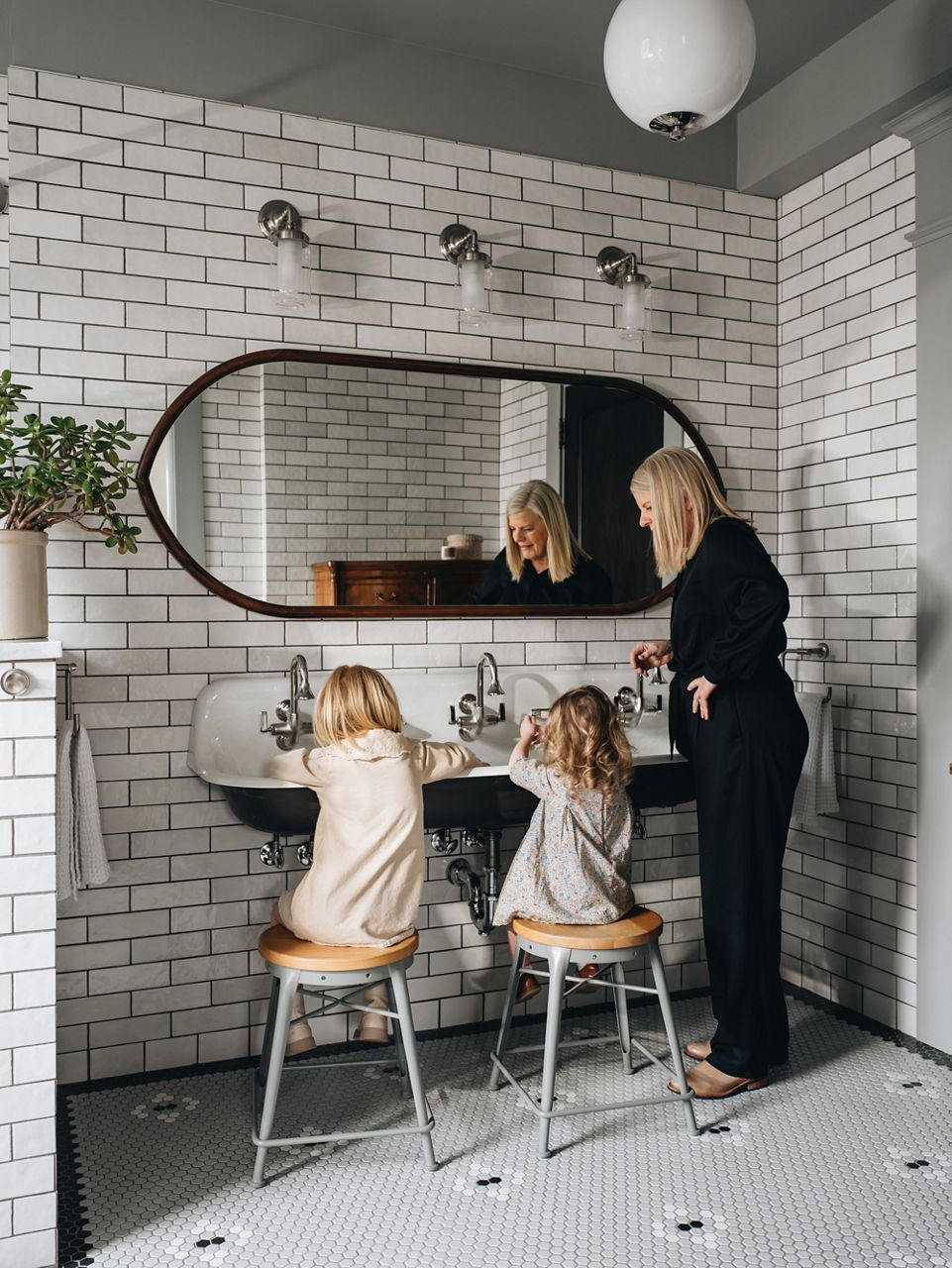 Designer Jean Stoffer and two small children using a trough style cast iron sink with three faucets in a bathroom with white subway tile.