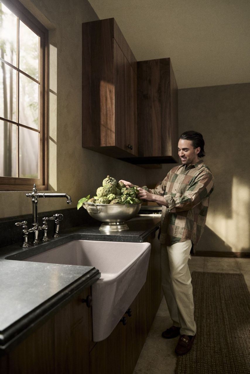 Jake Arnold touching a bowl of artichokes on the counter while standing next to a white farmhouse-style kitchen sink with a tall chrome faucet.