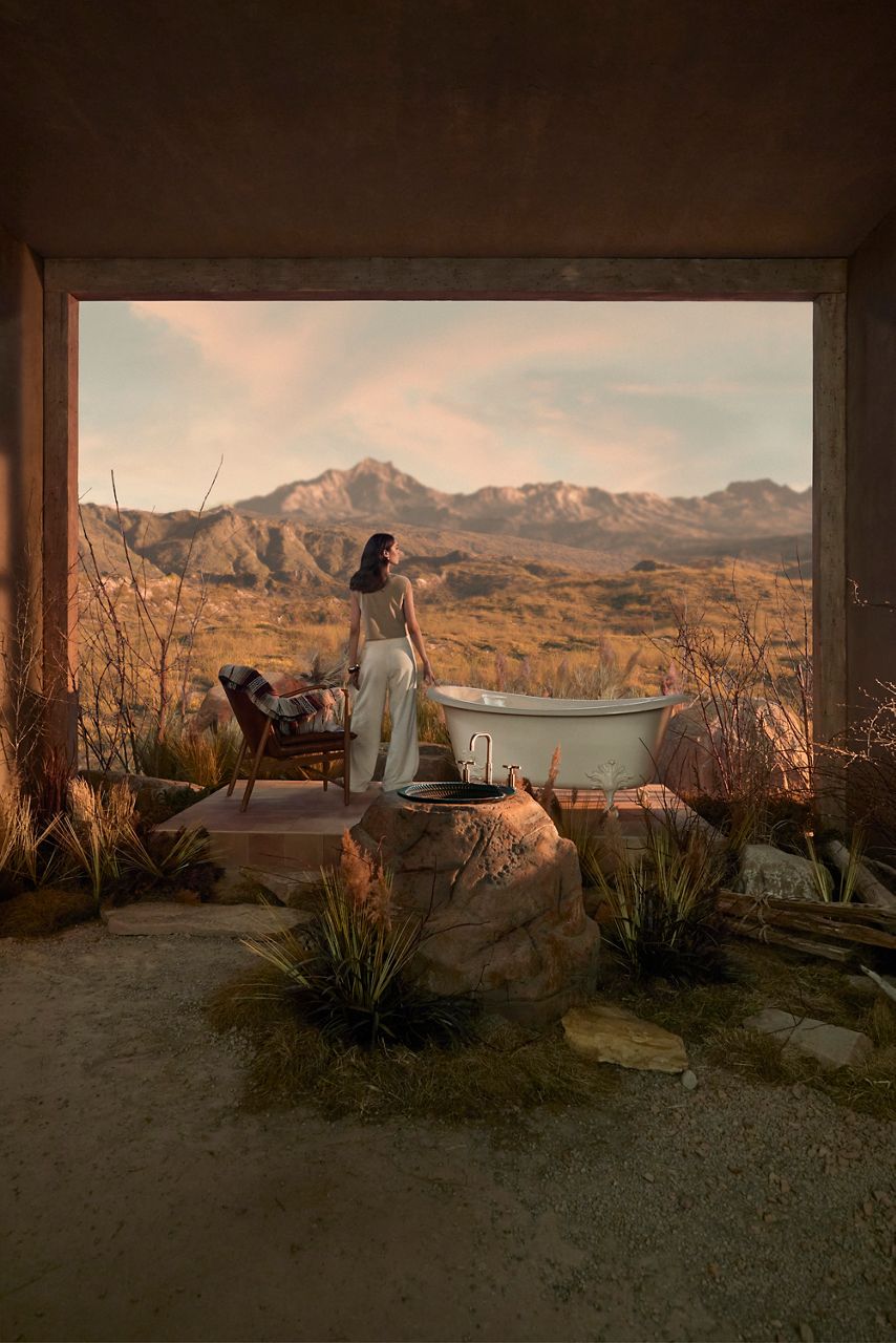 Outdoor bathtub scene set against a golden mountain landscape at sunset. A women in white pants and beige sleeveless top stands facing a freestanding clawfoot tub.
