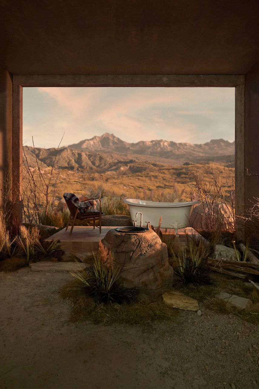 Scenic outdoor bathroom setting featuring a freestanding bathtub on a wooden deck overlooking a sunlit desert landscape with mountains, rocks, and natural vegetation in the background.