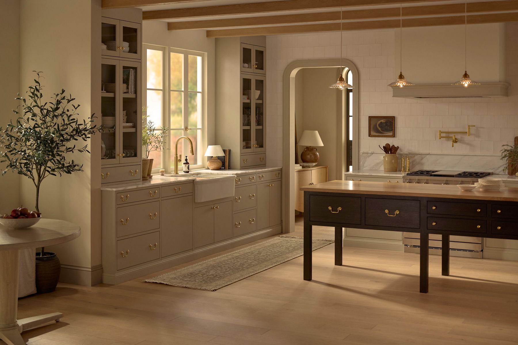 Warm, light-filled kitchen from the SMG Audrine Kitchen Collection featuring taupe cabinetry with brass hardware, a white farmhouse sink beneath tall windows, marble backsplash and countertops, exposed wood ceiling beams, and a dark wood island with classic drawer detailing.