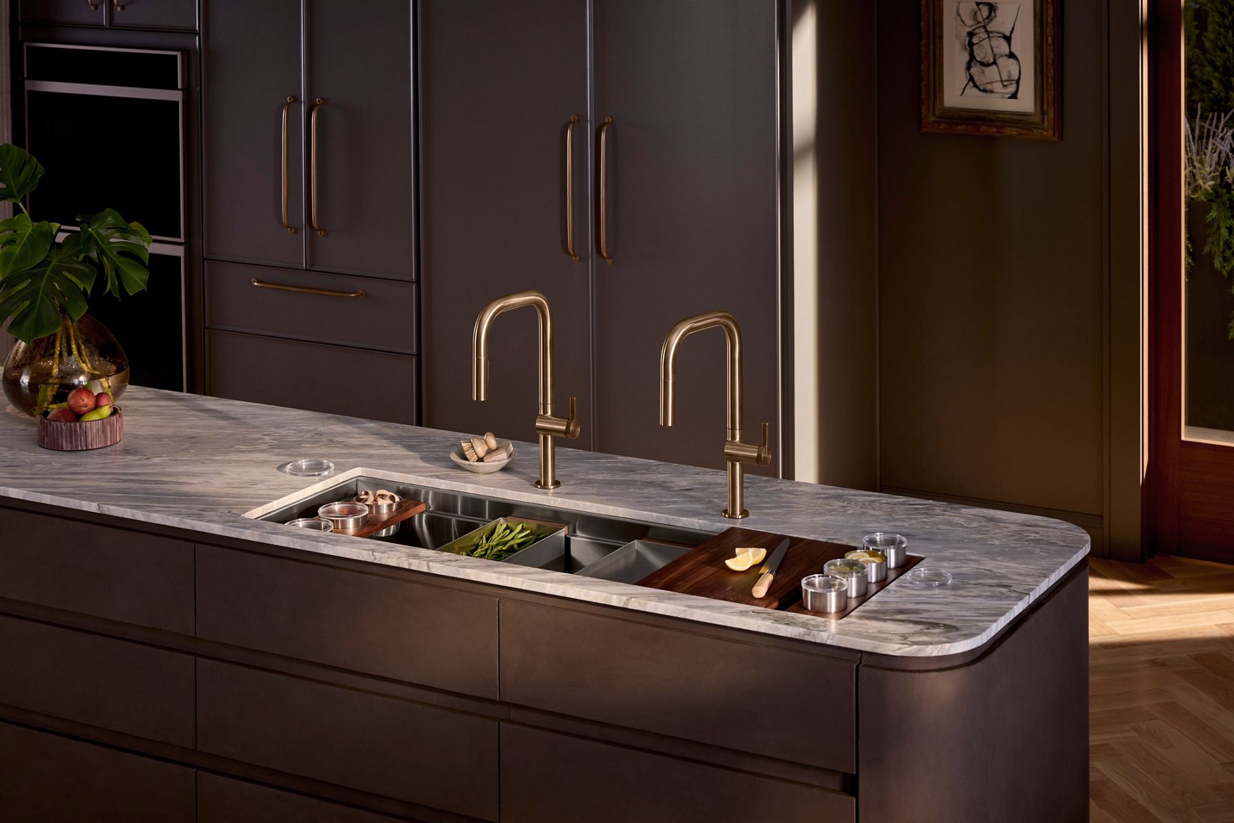 A modern, neutral-toned kitchen with two arched gold faucets, stainless steel sink, and marble countertop.