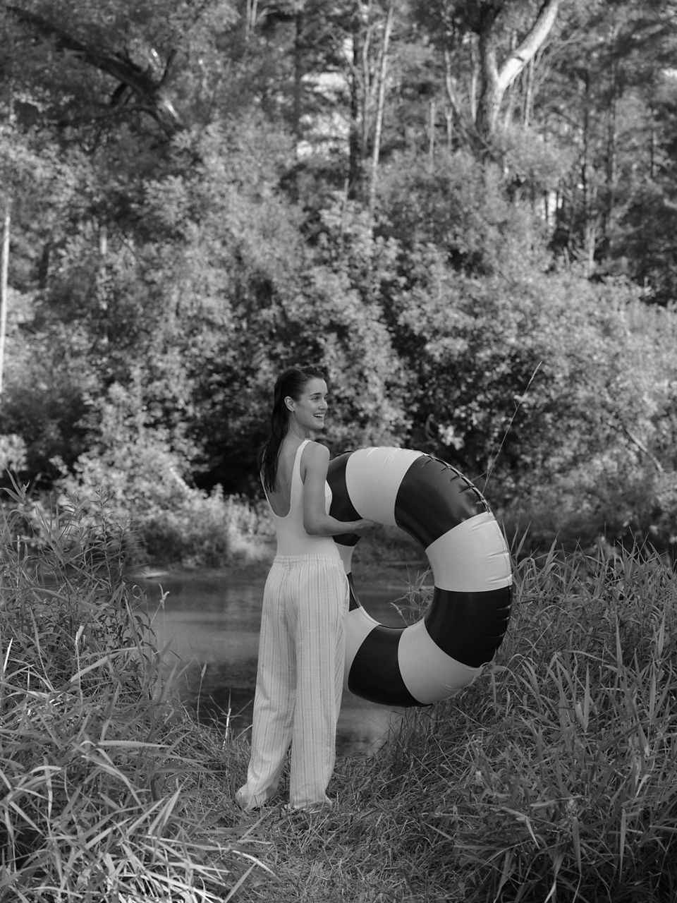 Black-and-white photo of a woman in a swimsuit holding a striped pool float with trees in the background.