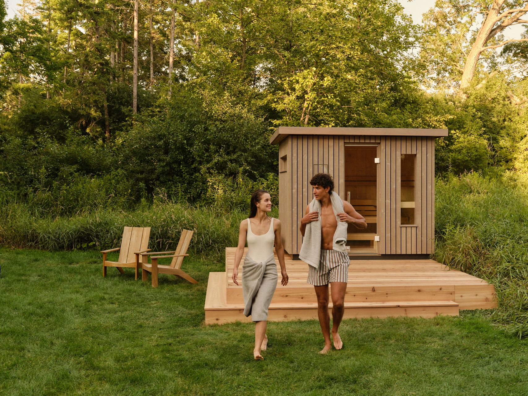 A modern wooden sauna set in nature, with two people smiling and walking outside on a deck.