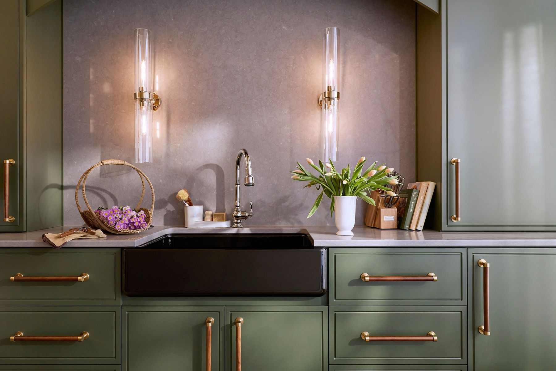 Black apron-front farmhouse sink with a polished chrome bridge faucet set against olive green cabinetry and brass hardware.