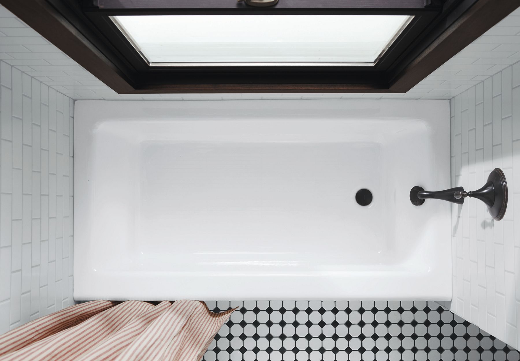 Glossy alcove bathtub shown in a bright bathroom with white subway tile, black and white flooring, and an orange striped shower curtain.