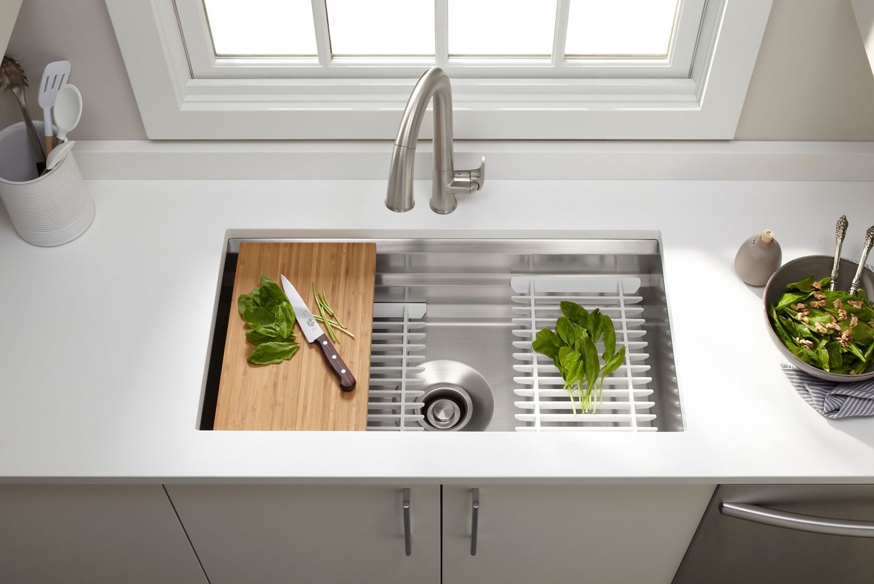 A workstation kitchen sink with a cutting board with a knife and de-stemmed spinach and other racks on its shelves.