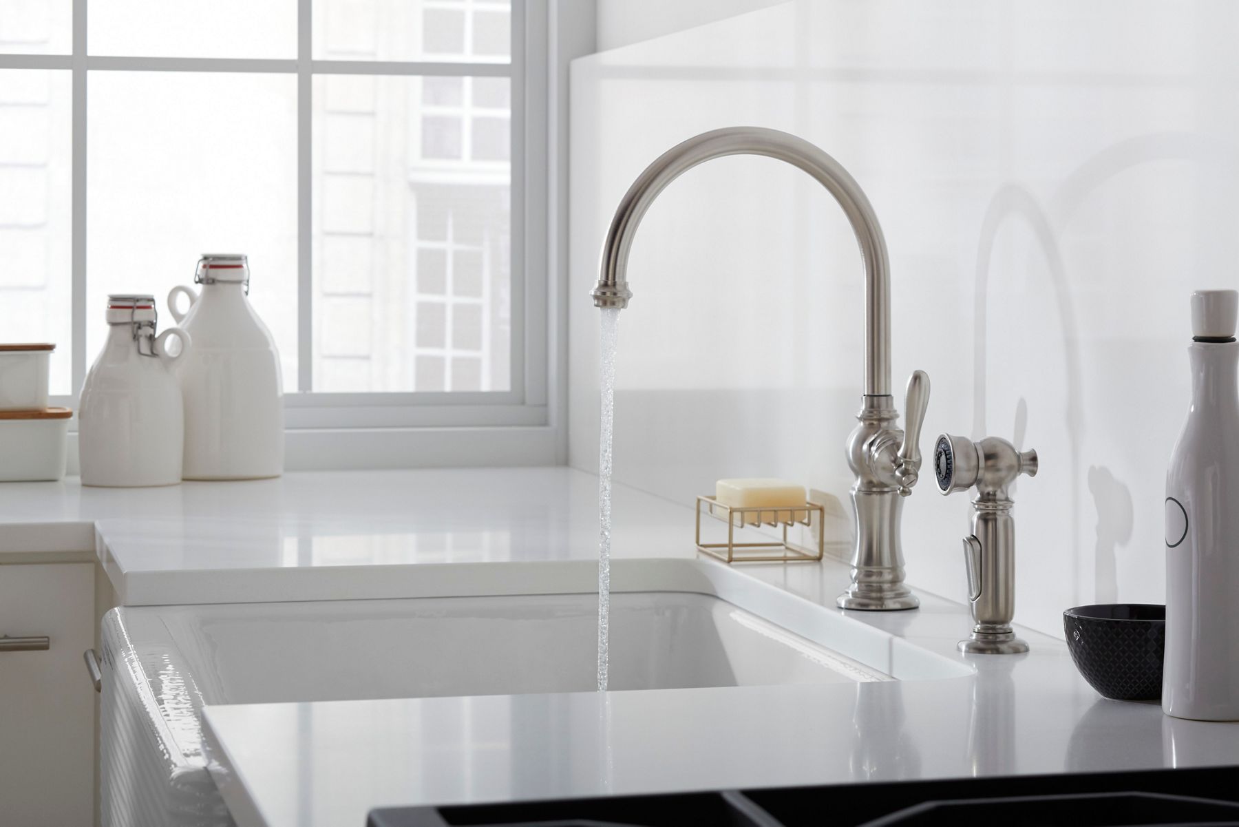 Modern white kitchen sink with a polished nickel faucet running water, styled with minimal countertop accessories and soft natural light from a large window.