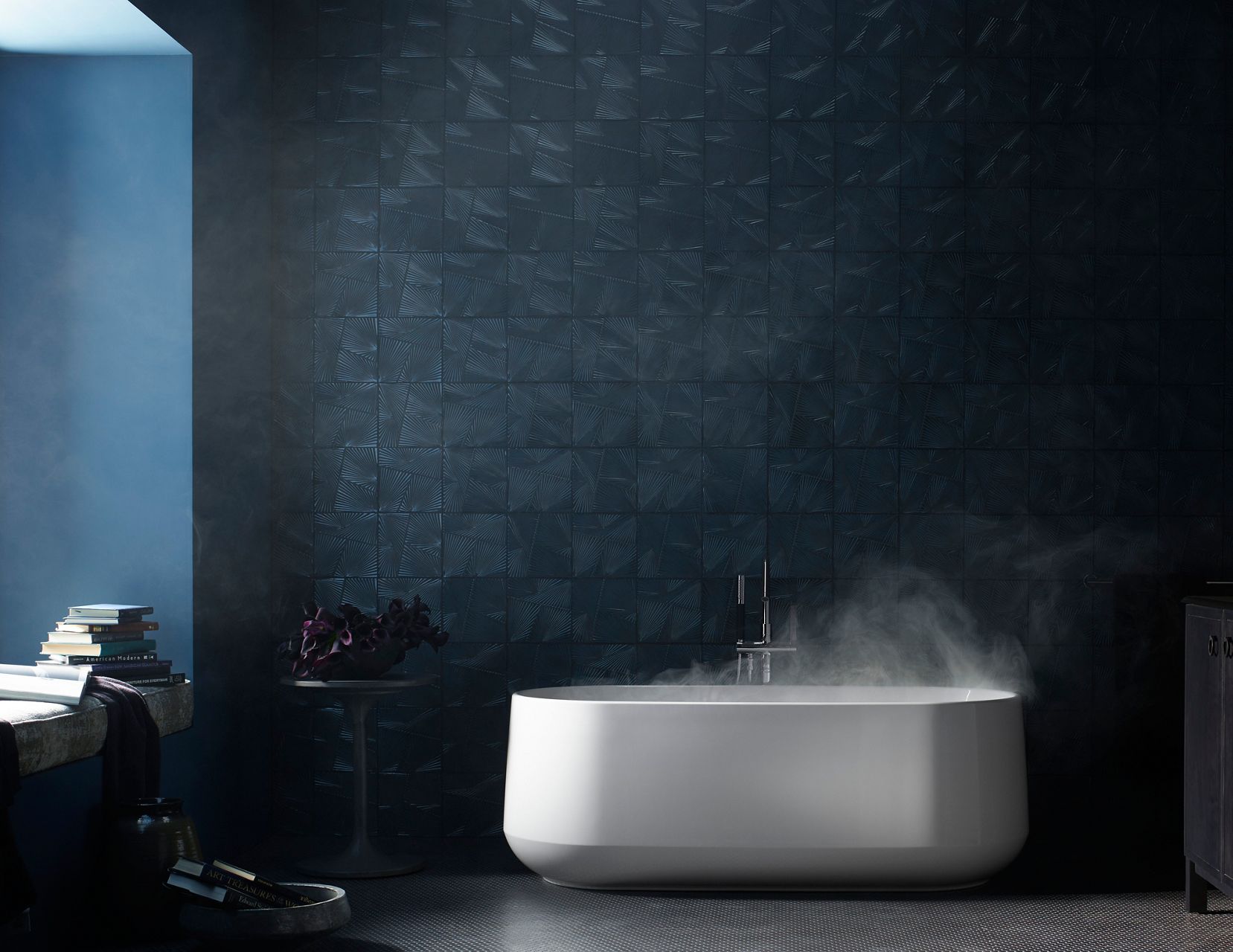 A white freestanding bath with steam rising from the surface in a bathroom with dark walls and floors, and light coming in on the left from an unseen window.