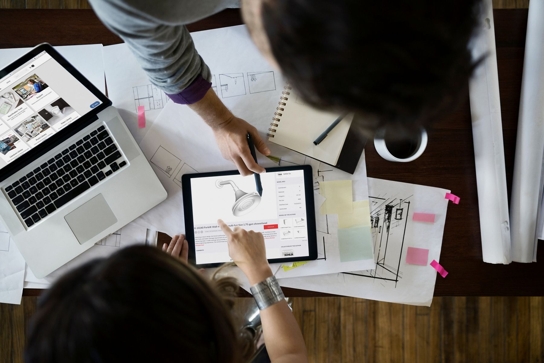 An overhead view of hands belonging to two people sitting across a desk from each other pointing to an image of a showerhead on a tablet with various other drawings and work materials scattered on the desk.