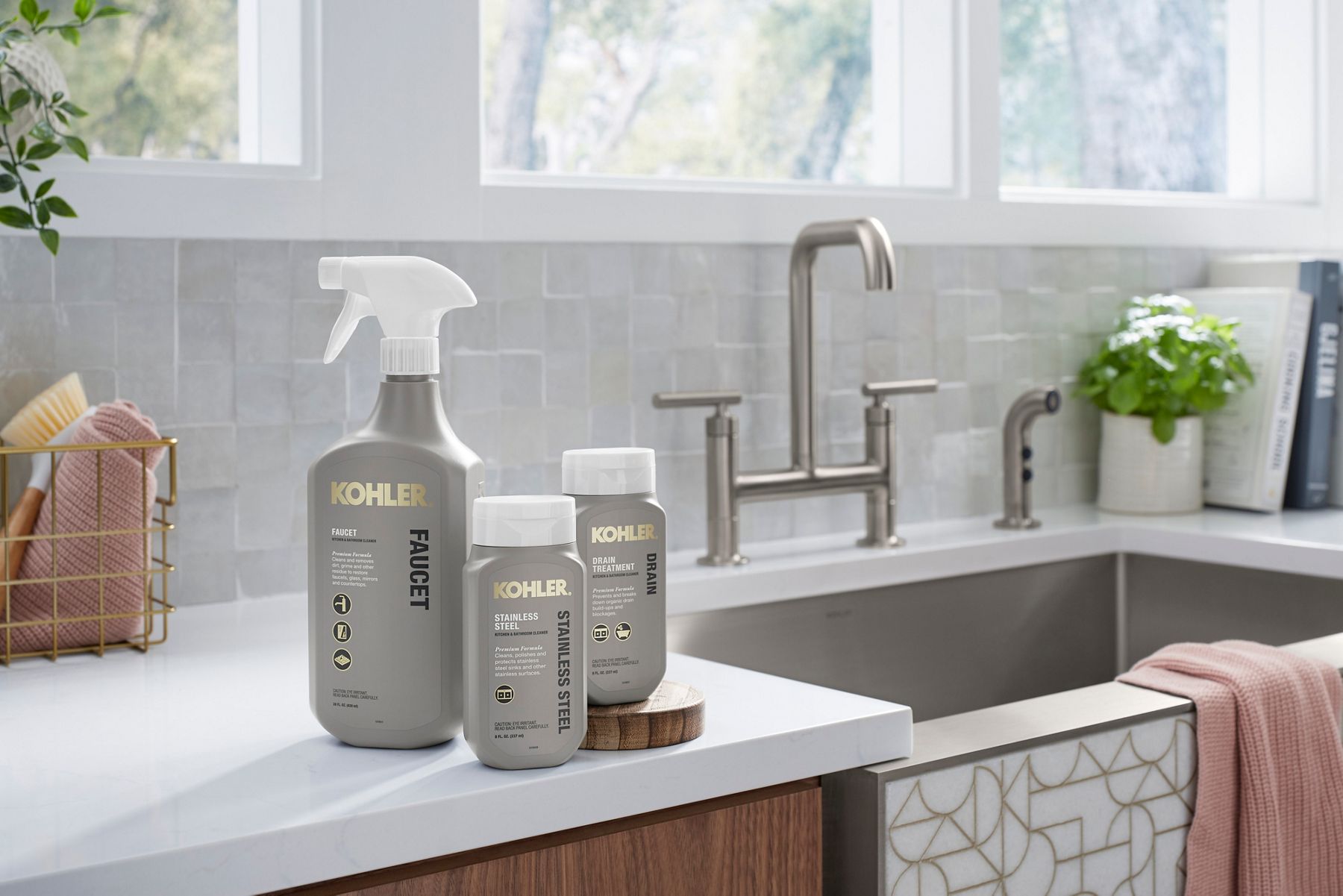 A spray bottle of Kohler faucet cleaner, a flip-top bottle of Kohler stainless steel cleaner, and flip-top bottle of Kohler drain treatment sit on a white counter with a stainless steel faucet and spray, stainless steel sink, backed gray tile backsplash. 