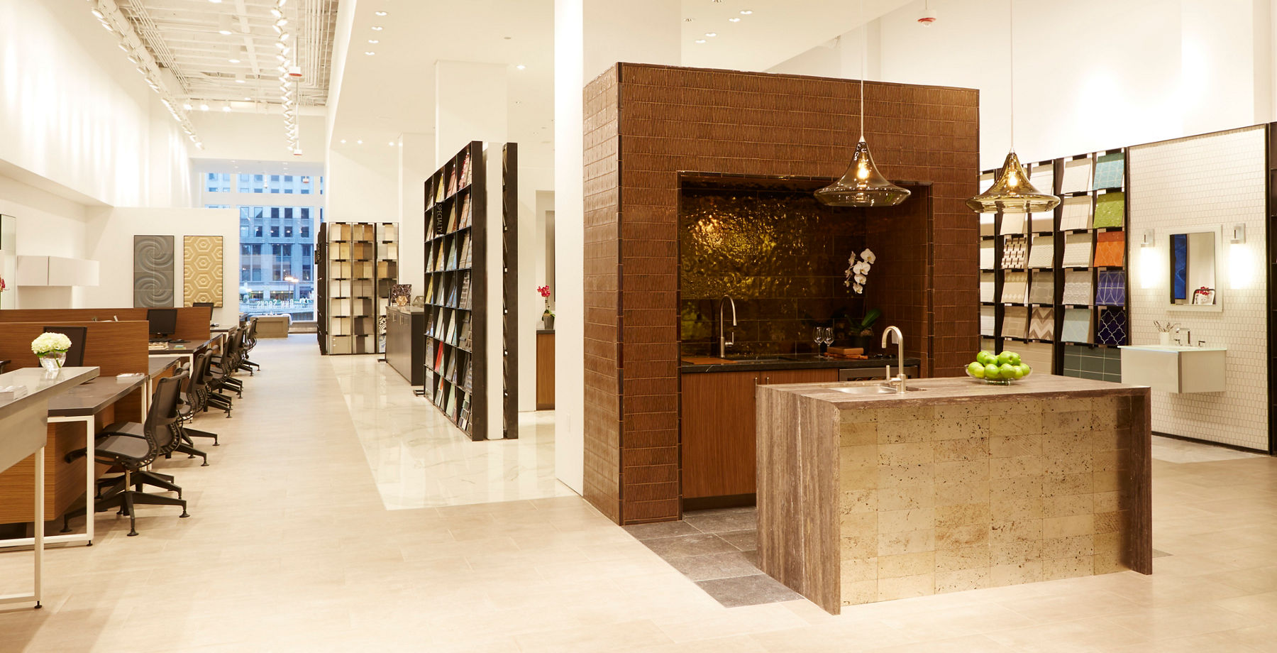 Modern interior design showroom featuring furniture and decor displays, a central island with pendant lights, sample shelves, and seating areas for client consultations.