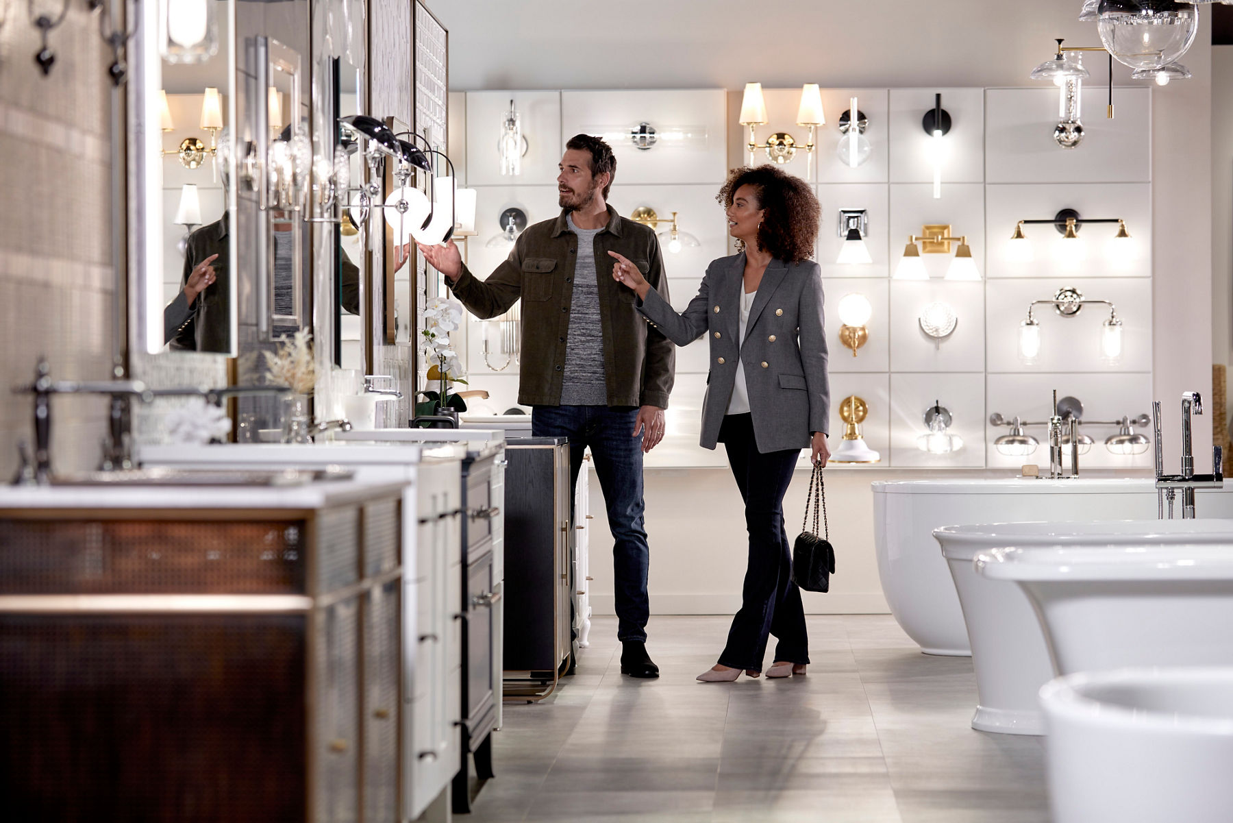 Wide image of a couple shopping inside of a bright and open Kohler store, looking at a wall of bathroom vanities, lights, and mirrors. They are pointing and reaching out to touch a lighting sconce. 