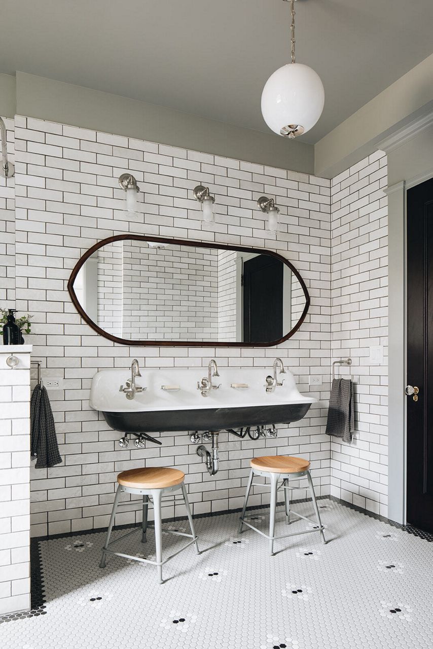 A modern black-and-white bathroom with a black-framed round mirror, double white sink, and wood stools, illustrating a sustainable design.