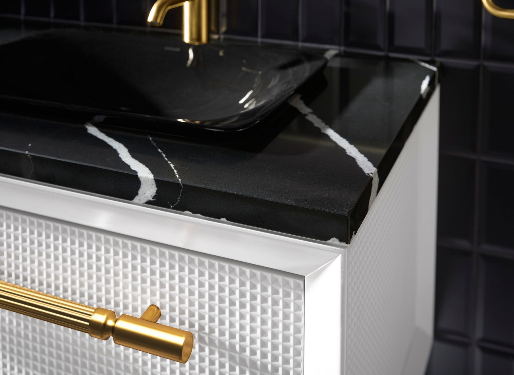 Close up of a white bathroom vanity, showing the waffle-patterned texture of the drawer, the black stone counter top with white veining, and the brass accents and faucet.