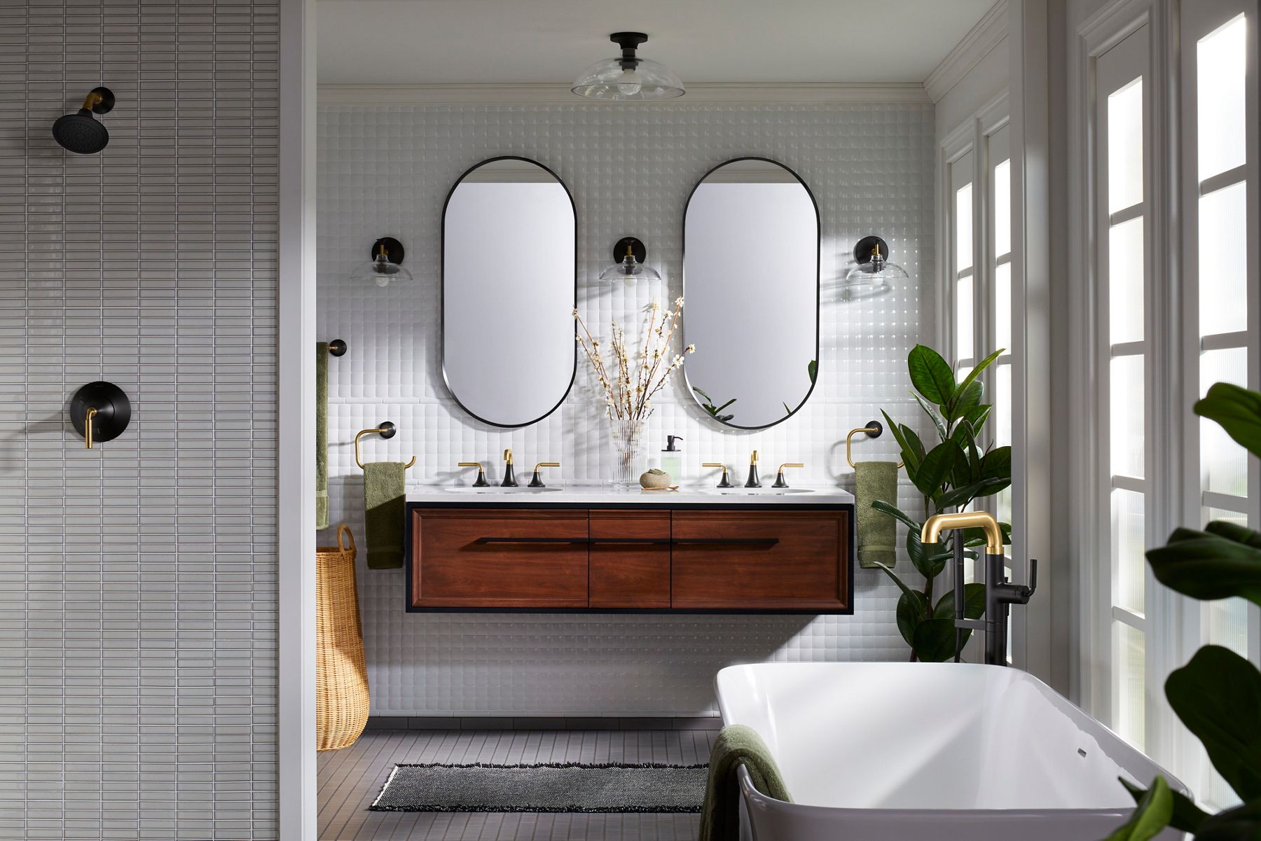 A bathroom styling space with a wall-mount double vanity in wood tones with black accents and two-tone faucets on a white countertop, with a white tile wall, two pill-shaped mirrors and sconce lights.