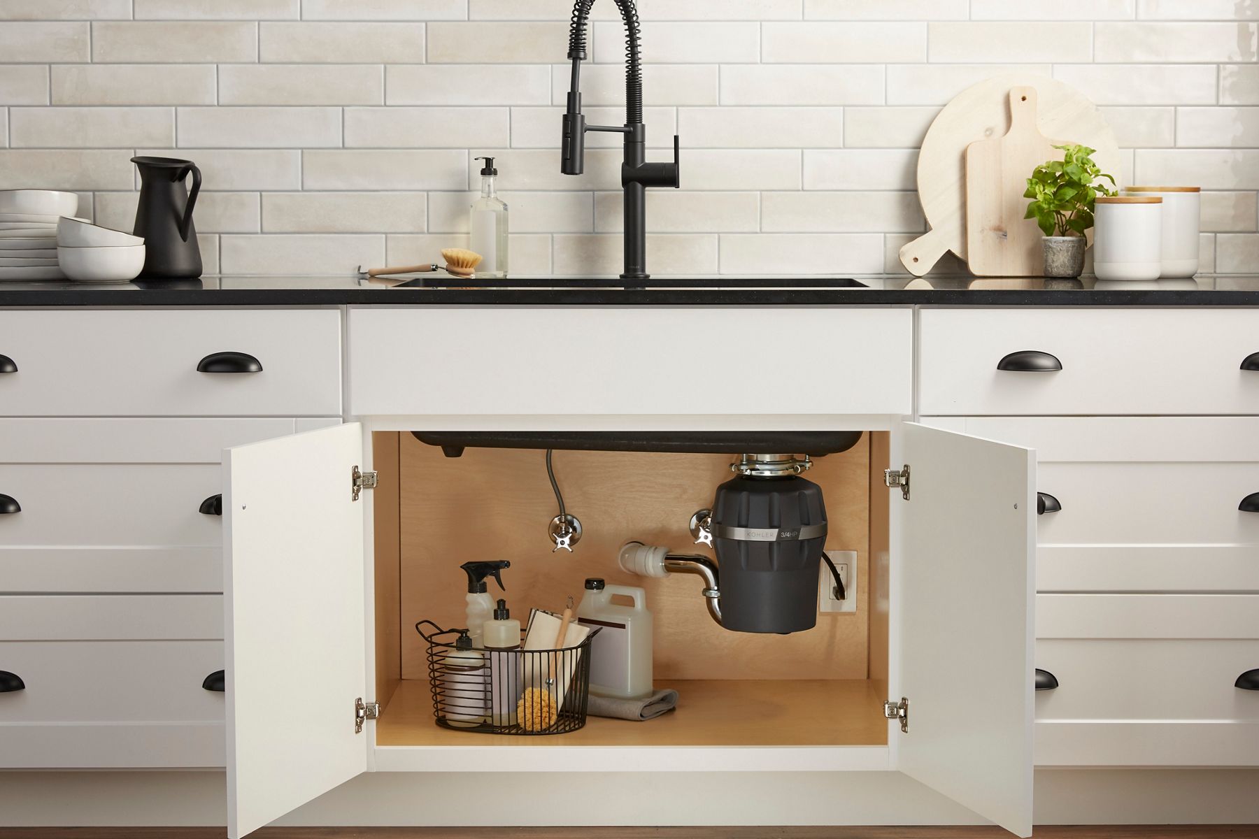 A white kitchen cabinet with the doors under the sink open to reveal a garbage disposal and various kitchen cleaning items.