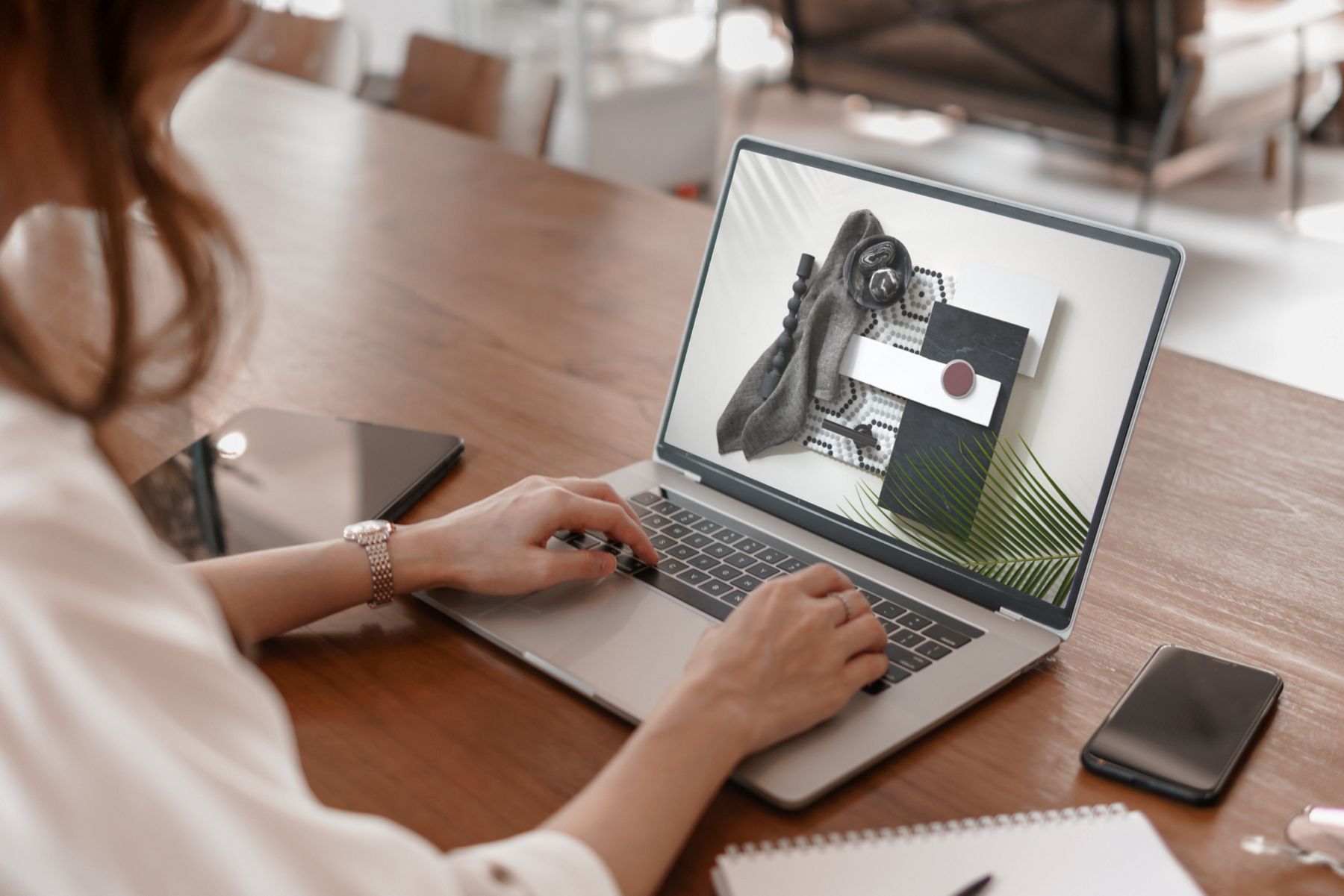 Two hands, at a table, typing on an open laptop with design samples on screen.