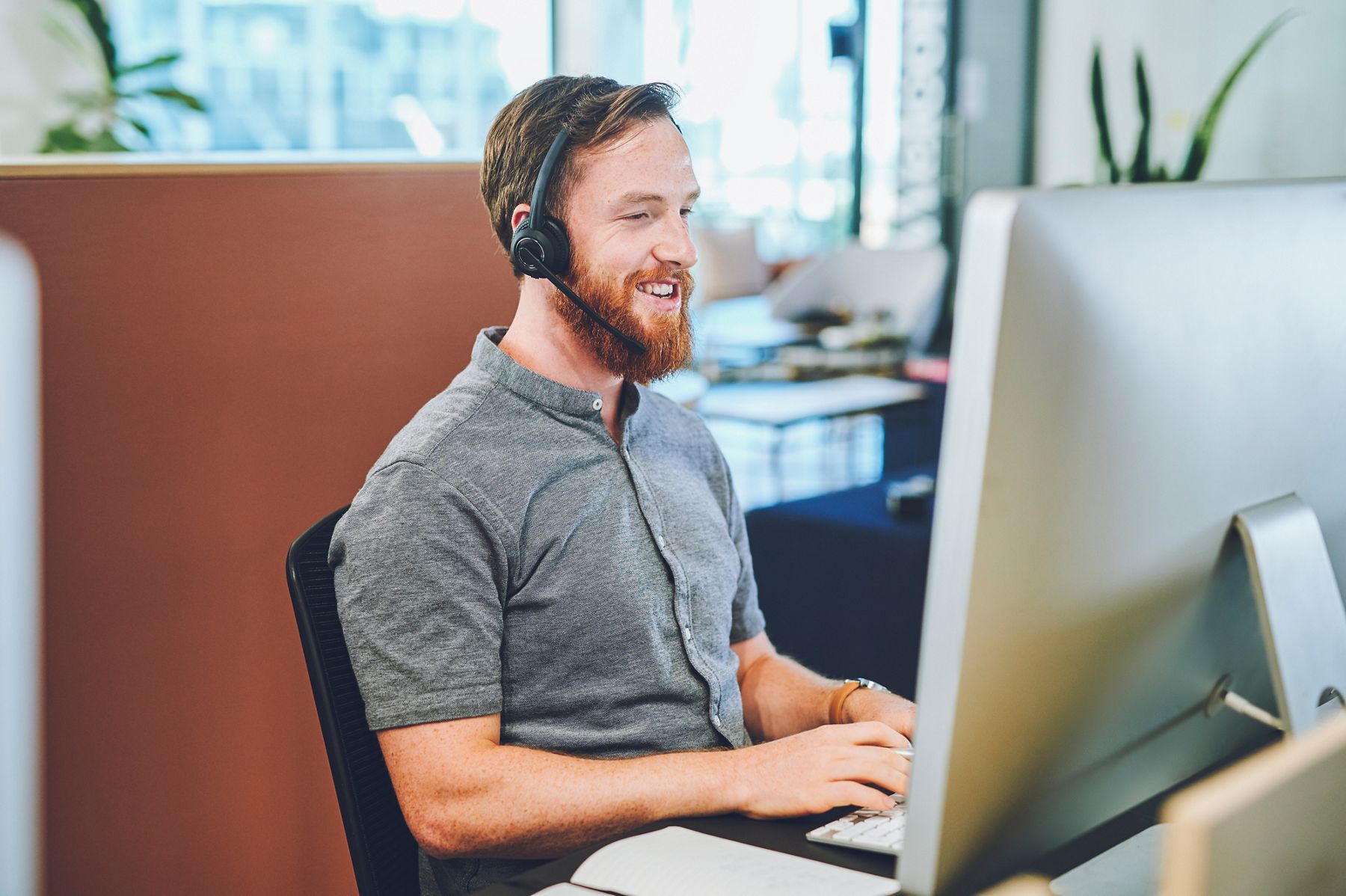 A similing man with a beard wearing a headset and short sleeve shirt sits at a desk in front of a computer.