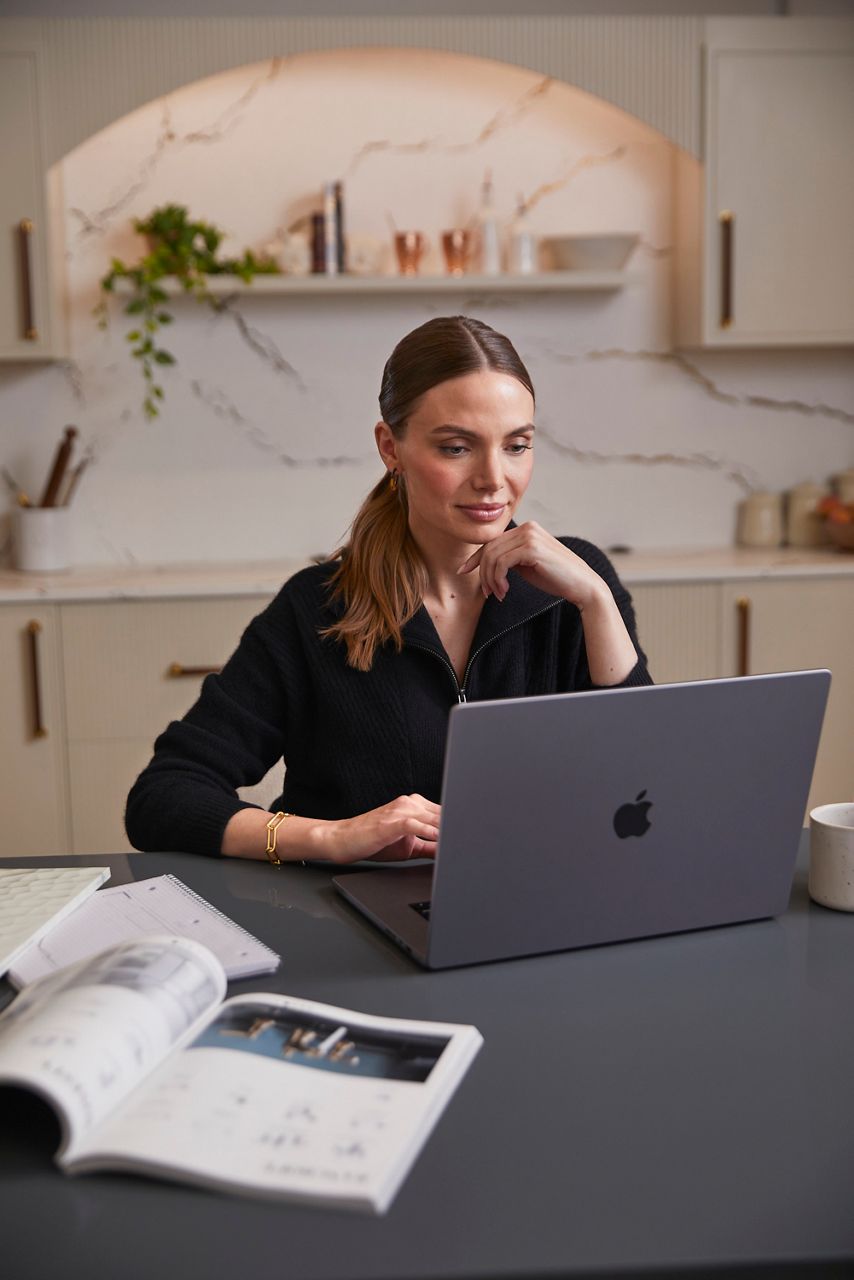 A woman sitting at a kitchen countertop in front of an open laptop with an open catalog in the foreground, backed by marble wall, shelf, and cabinets.