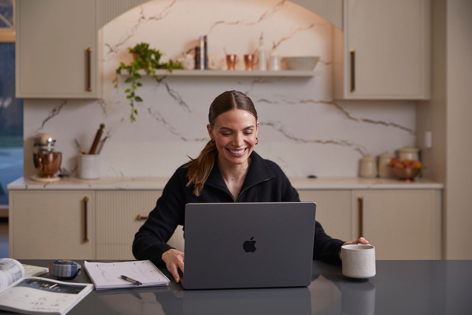 A similing woman siitting at a kitchen countertop in front of an open laptop and holding a mug, backed by marble wall, shelf, and cabinets.